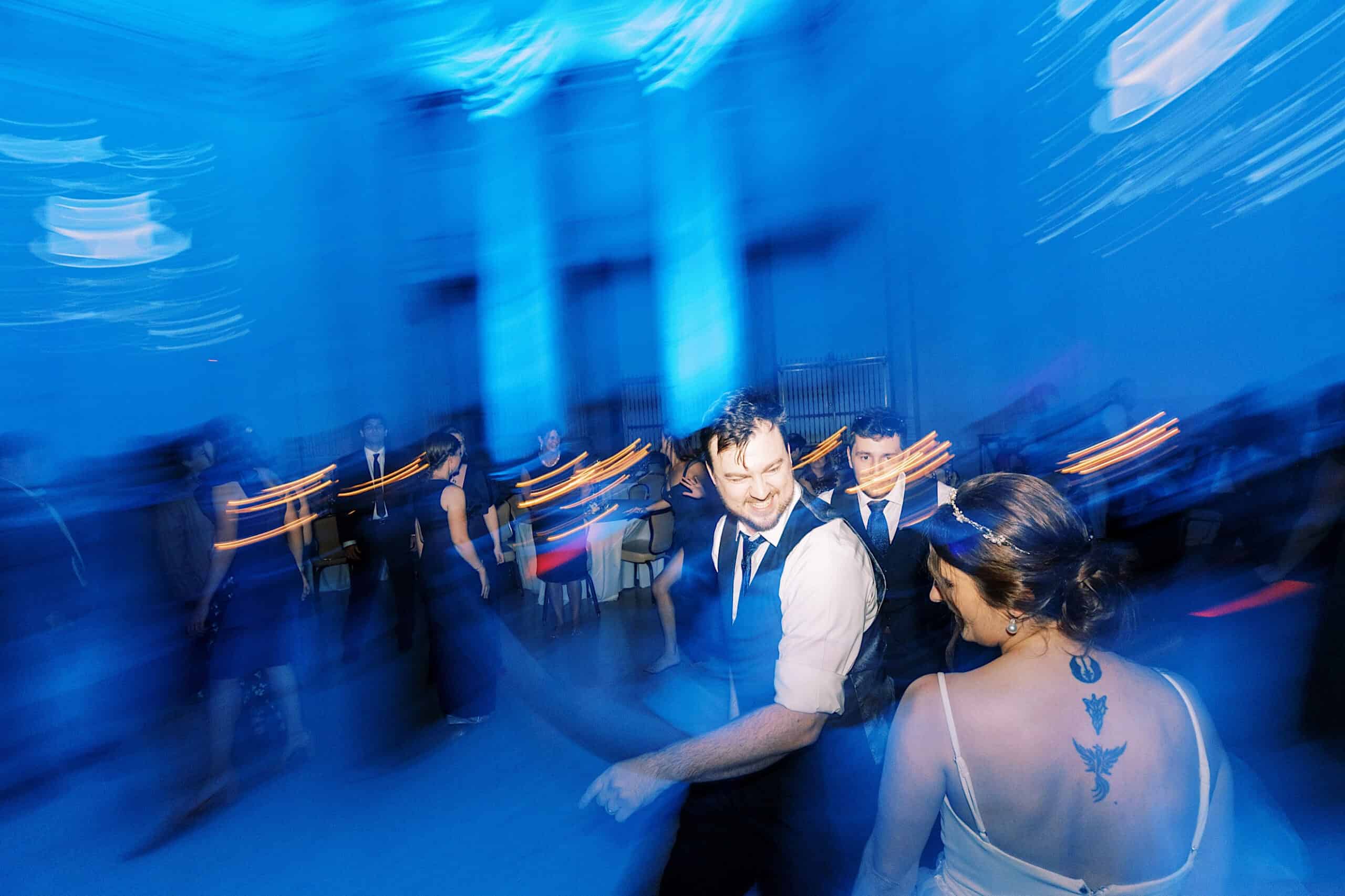 People in formal attire dance in a blue-lit room at a Franklin Institute wedding; motion blur adds energy, and a woman with a tattoo stands in the foreground facing a smiling man.