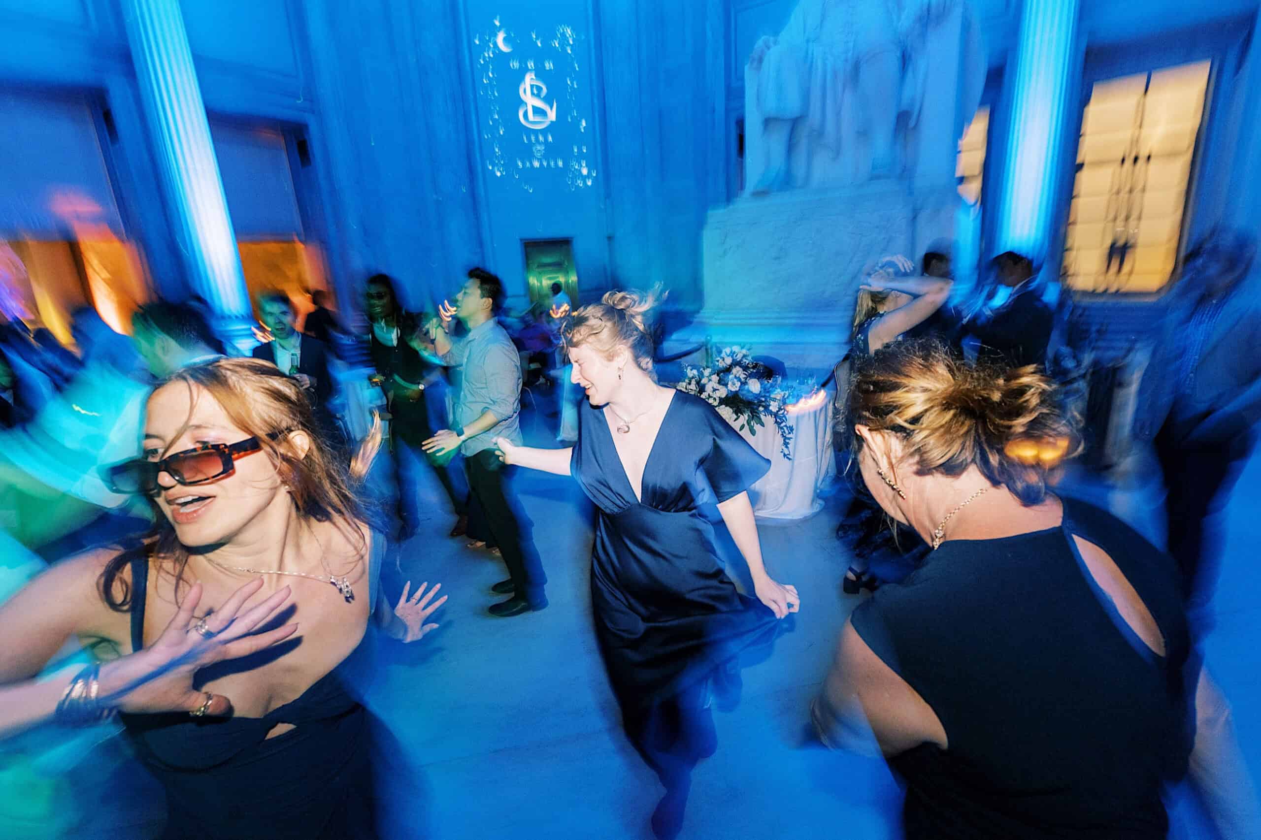 People dance energetically in a blue-lit room with columns and statues at a Franklin Institute wedding, wearing formal attire. The image has motion blur, capturing lively movement and a festive atmosphere.