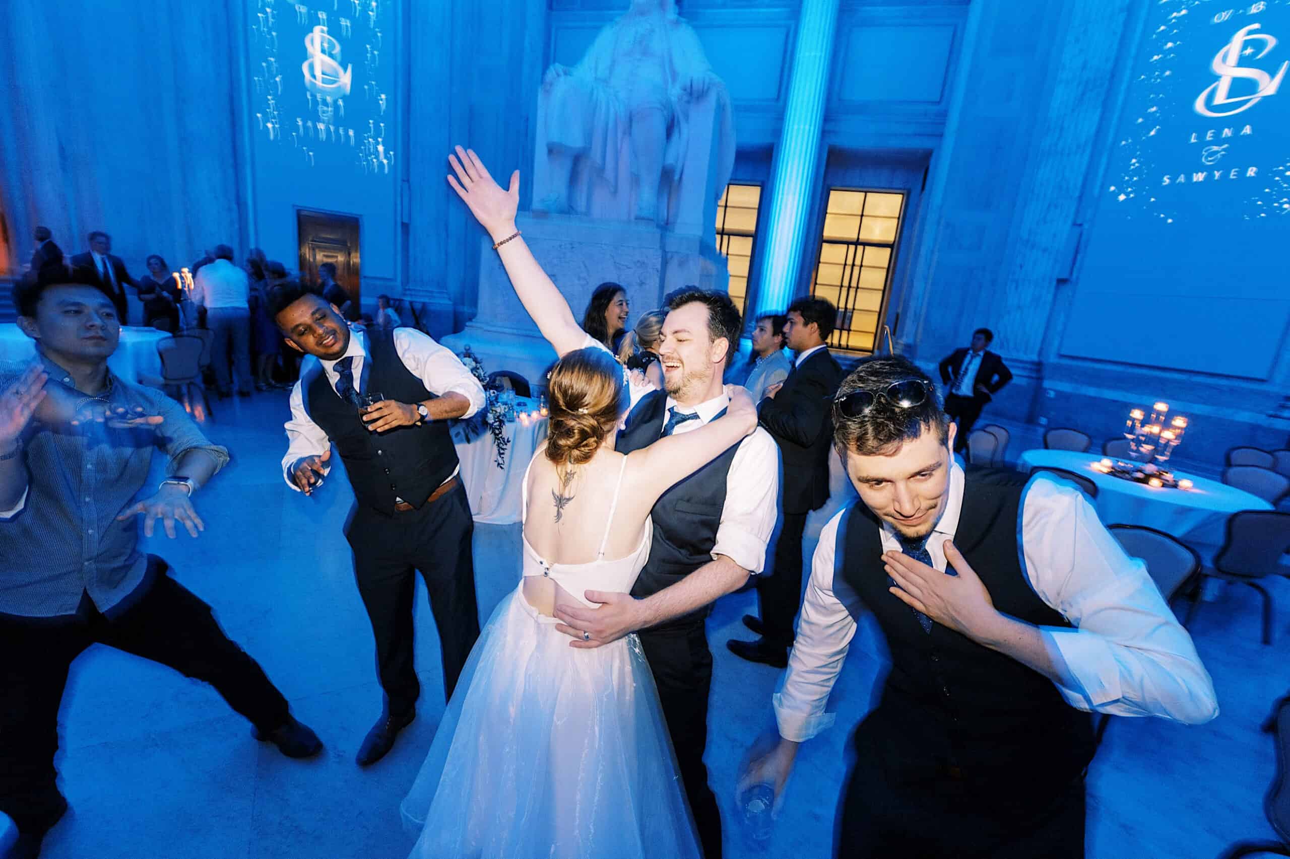 A bride and groom dance and embrace on a blue-lit ballroom floor at their Franklin Institute wedding, surrounded by friends in formal attire enjoying the celebration.
