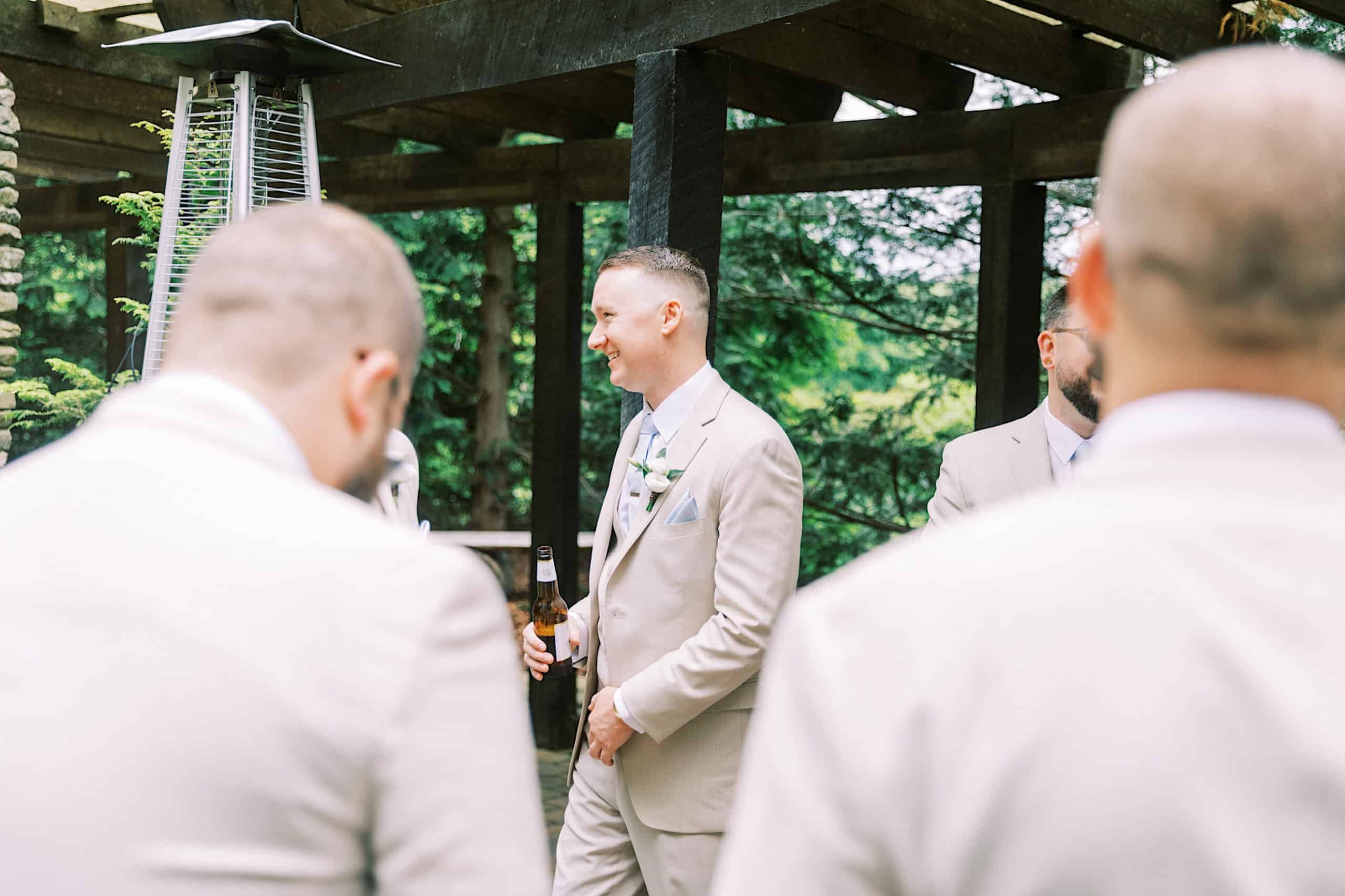 A man in a light-colored suit holding a beer bottle stands outdoors with other men in similar attire, surrounded by wooden beams and greenery.