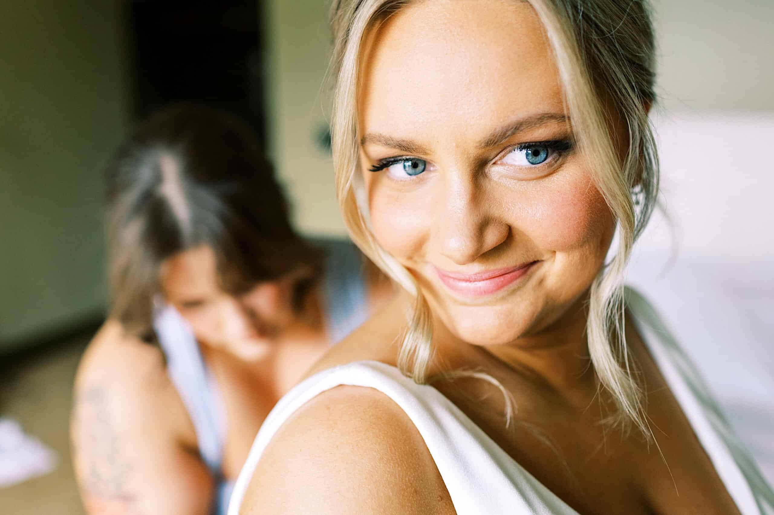A woman in a white dress smiles at the camera while another person helps adjust her dress in the background.