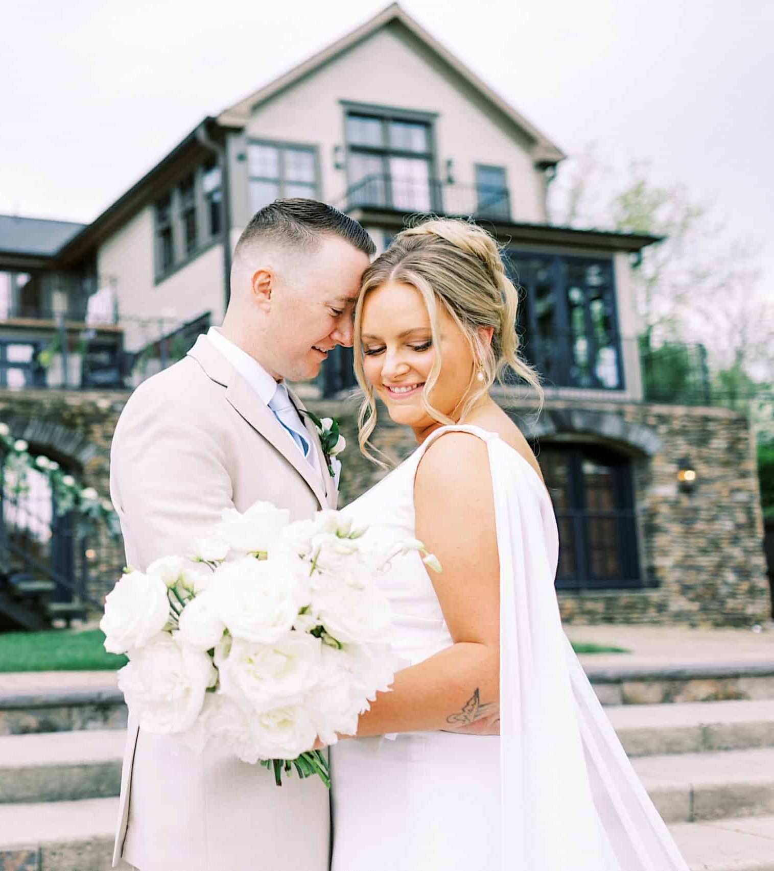 A bride and groom stand together outdoors, smiling and holding a white bouquet, with the elegant Lake House Inn Wedding venue’s stone house and decorated stairs in the background.