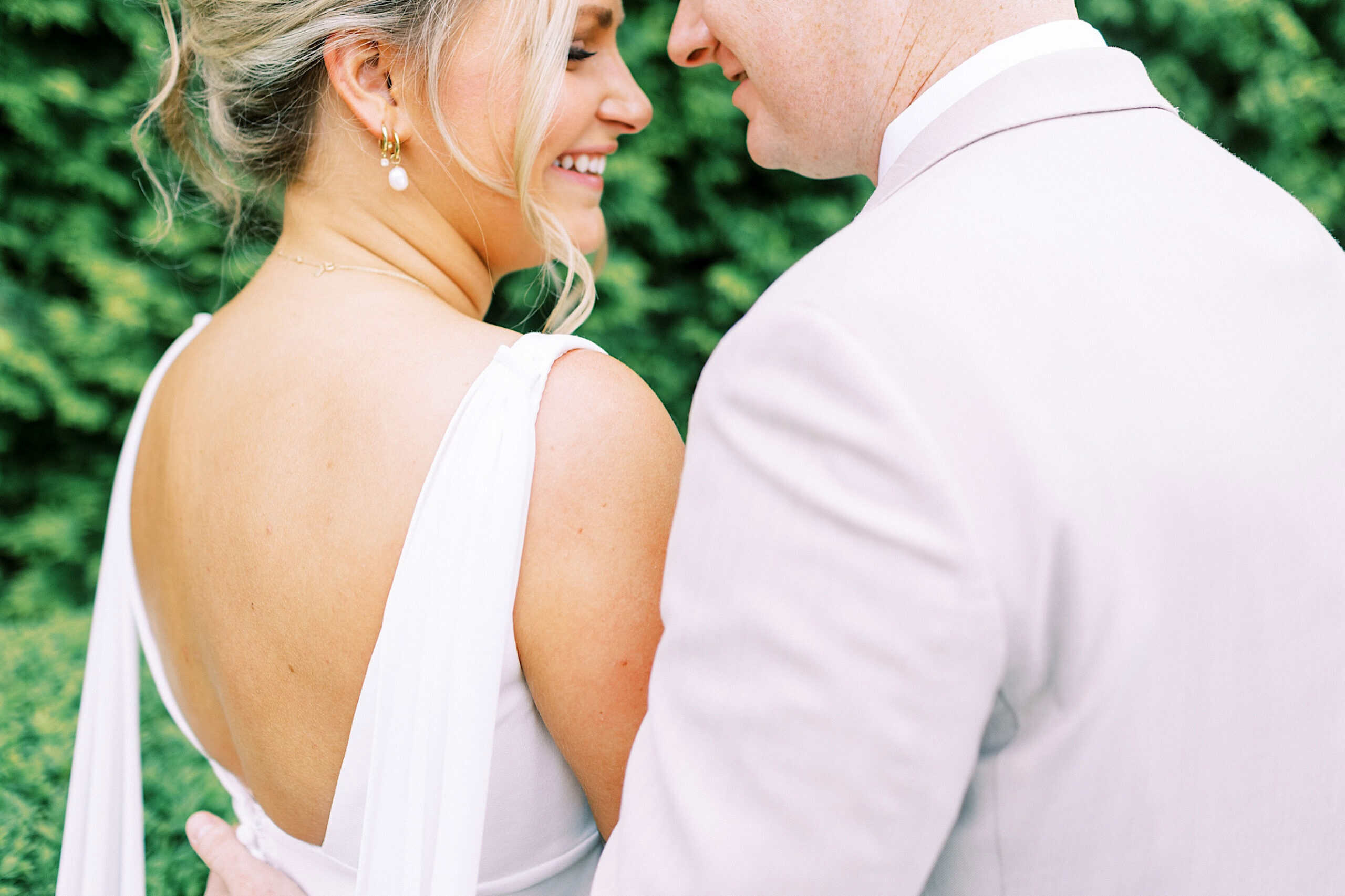 A couple dressed in light-colored formal attire stands close together outdoors, smiling, with greenery in the background.