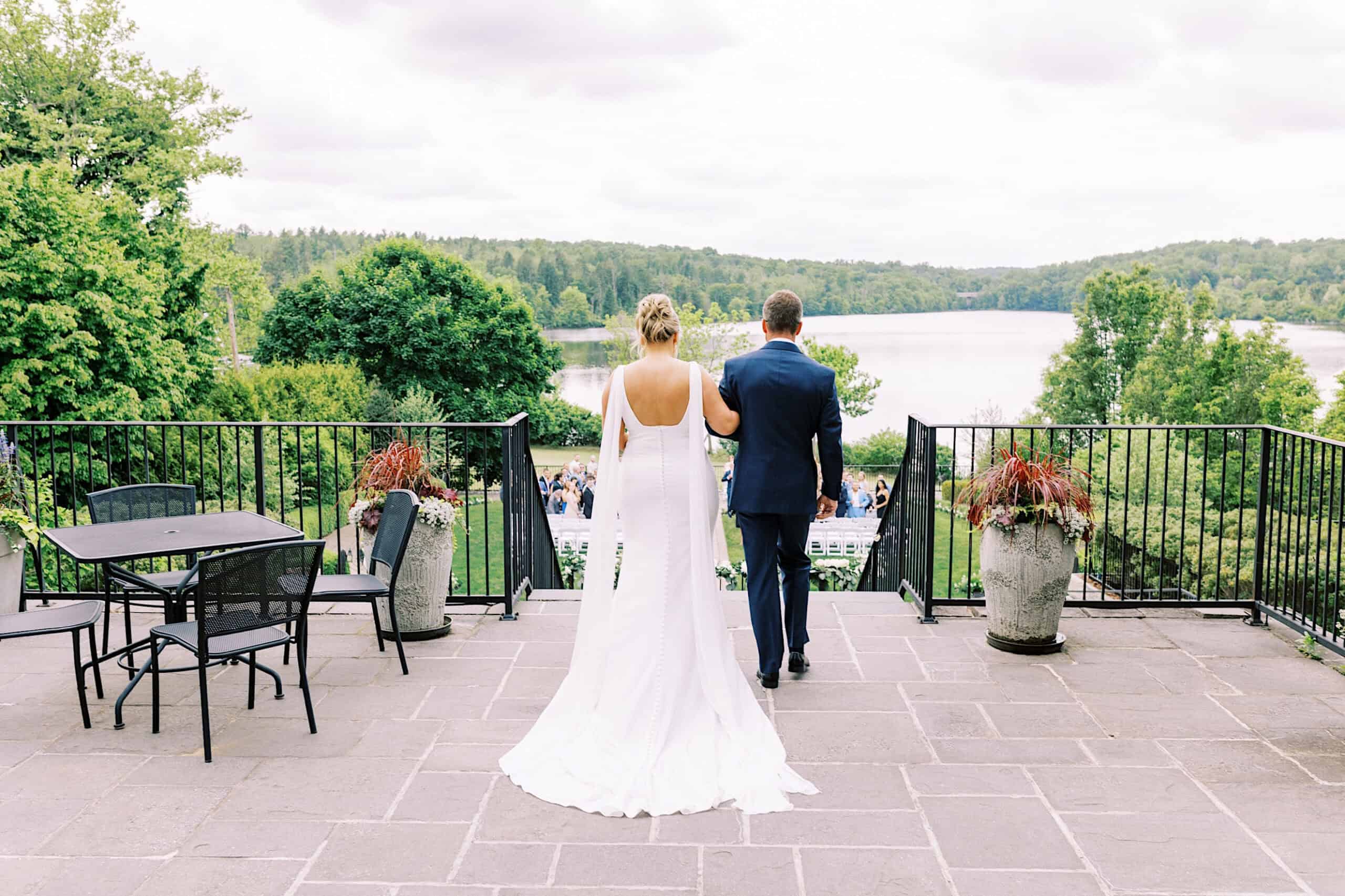 A bride in a white dress and a man in a suit walk arm-in-arm down a stone terrace at their Lake House Inn wedding, heading toward an outdoor ceremony overlooking the lake and trees.