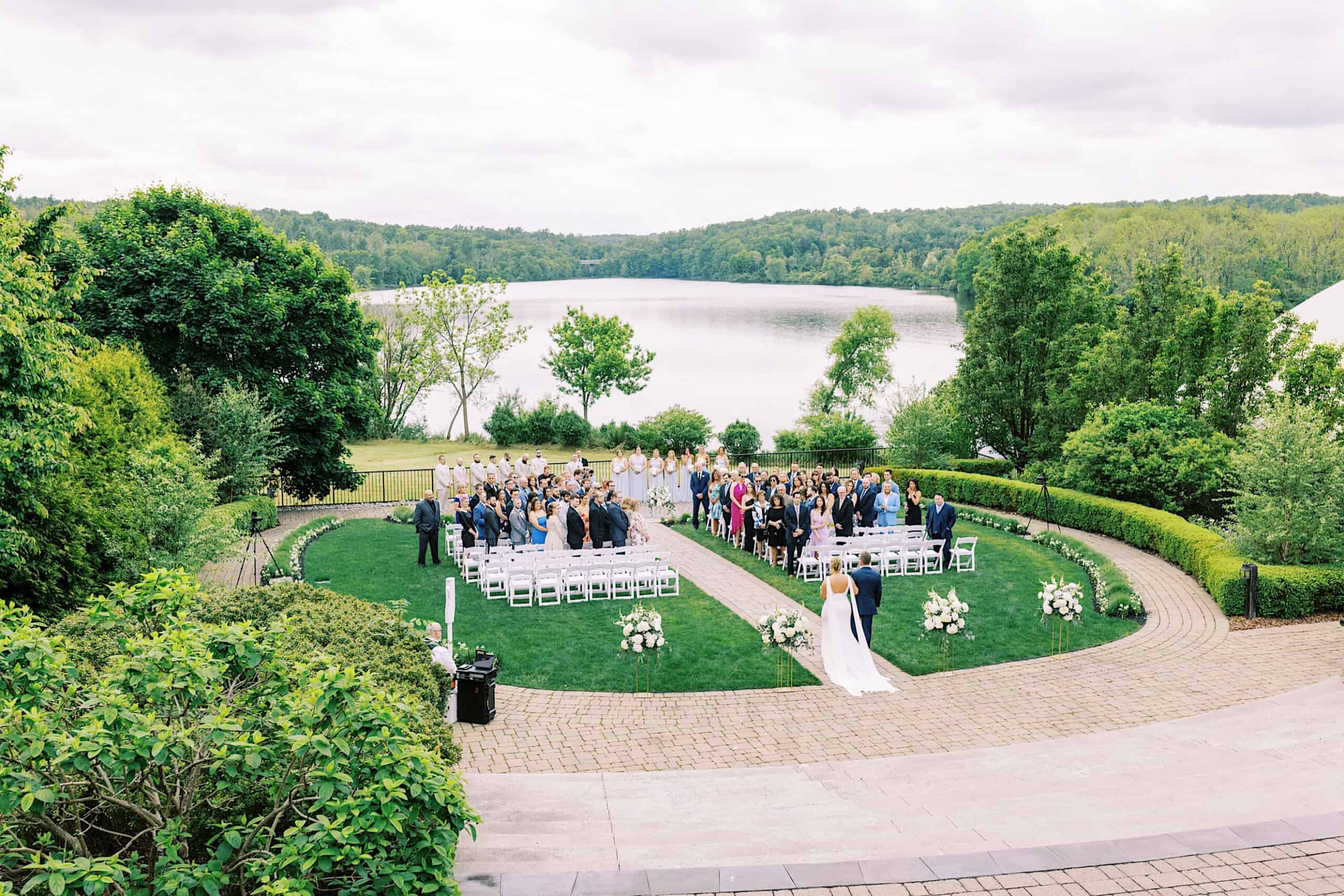 Outdoor Lake House Inn Wedding ceremony by a lake, with guests seated on white chairs in a semicircle, a bride walking down the aisle, and lush greenery surrounding the area.