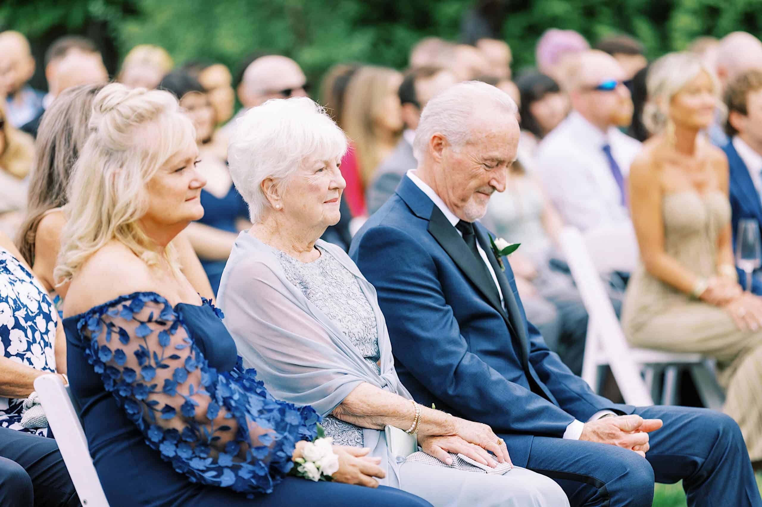 People sit in rows of white chairs at an outdoor Lake House Inn Wedding, with two women and a man in the foreground dressed in formal attire, watching attentively.
