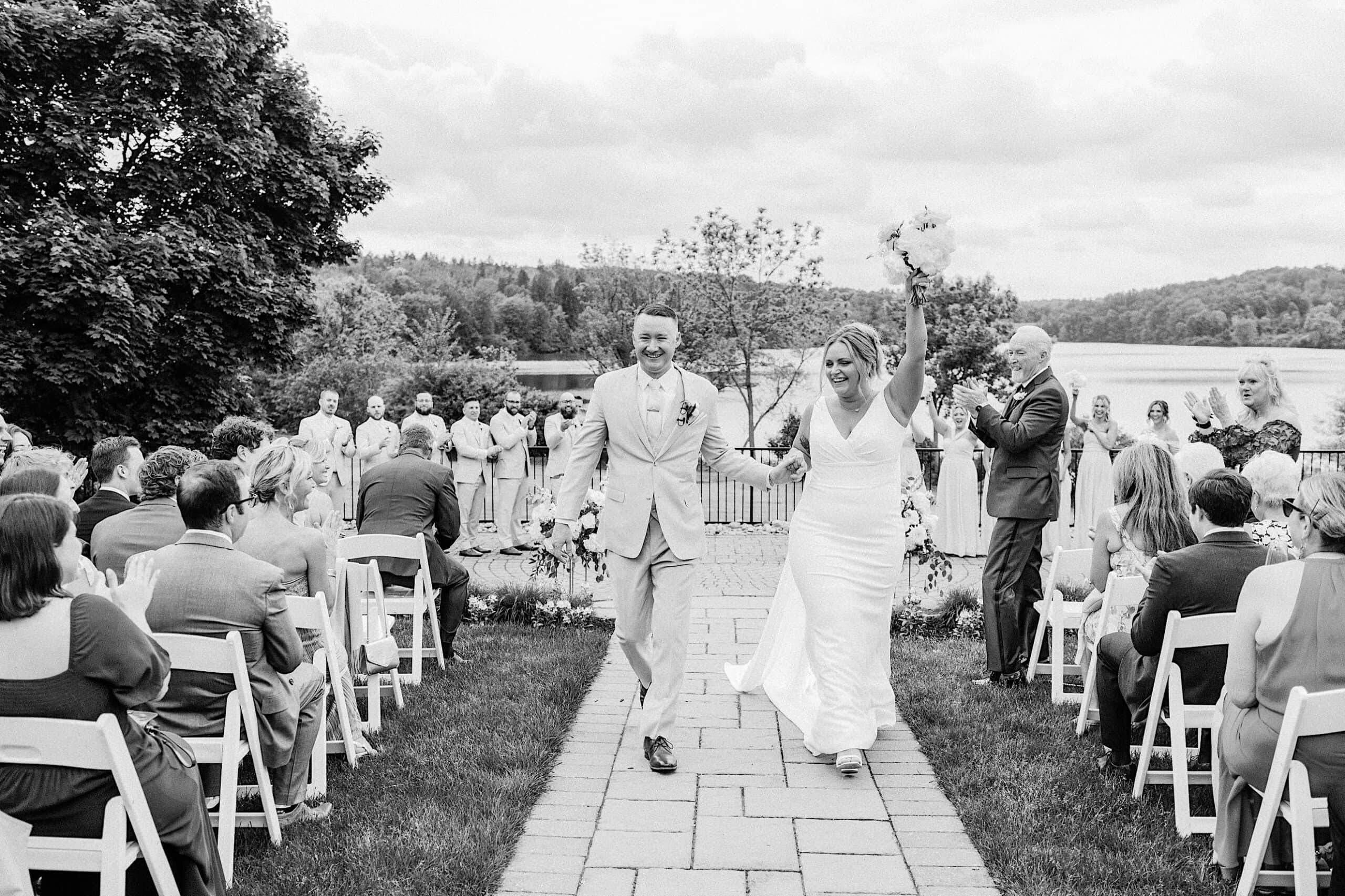 A bride and groom walk down an outdoor aisle at their Lake House Inn Wedding, smiling and holding hands, while guests seated on both sides applaud.