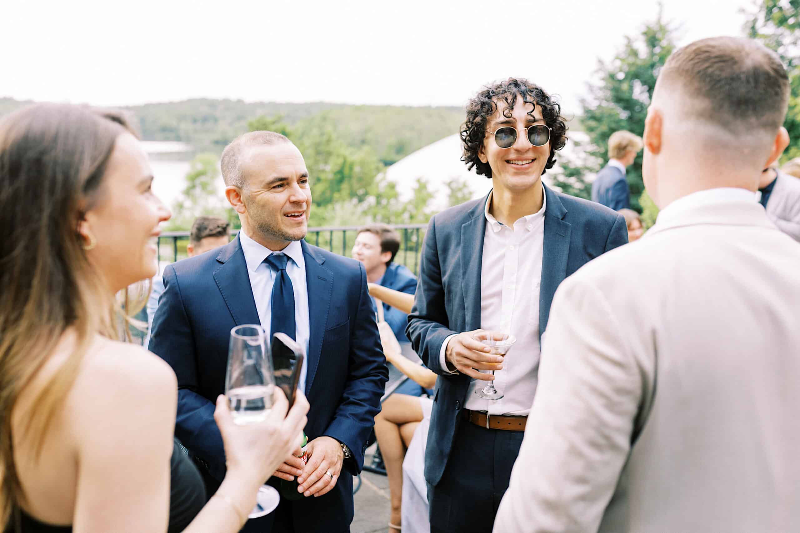 Four people in formal attire stand outdoors at a Lake House Inn wedding, holding drinks and talking, with others and lush greenery visible in the background.