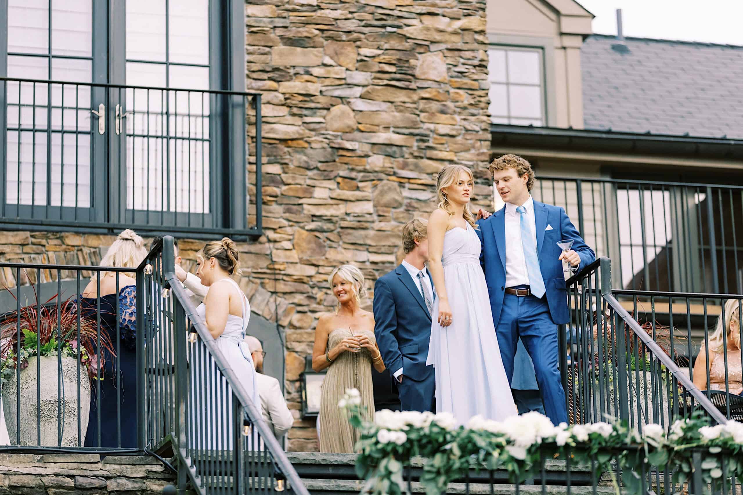 A group of people dressed formally walk down outdoor stairs next to a stone building, surrounded by flowers and greenery at a picturesque Lake House Inn wedding.