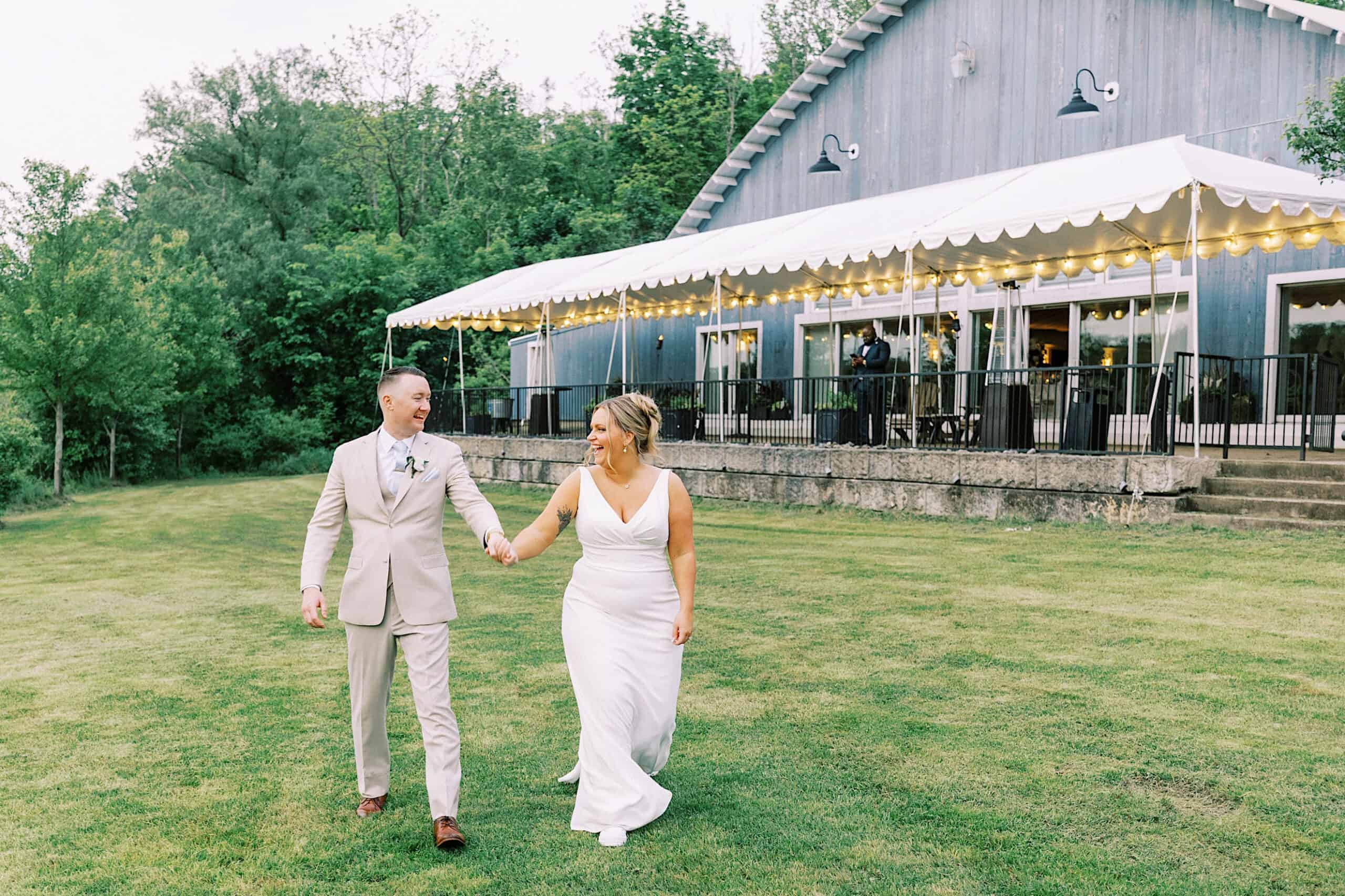 A bride and groom walk hand in hand on a lawn outside a barn-style event venue decorated with string lights, capturing the rustic romance of a Lake House Inn Wedding.
