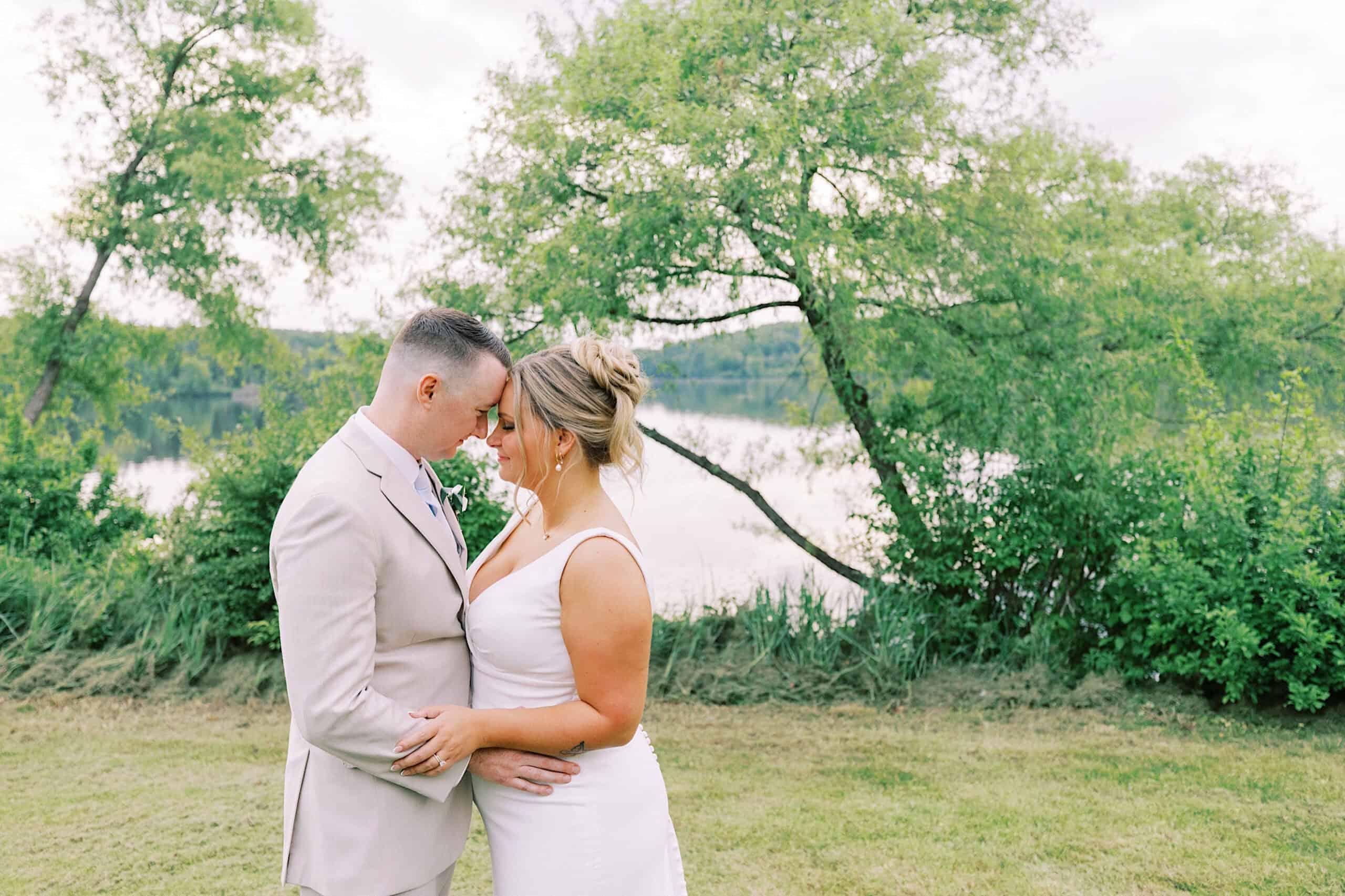 A couple in wedding attire stands forehead to forehead outdoors near greenery and a lake at their Lake House Inn Wedding, holding each other closely.