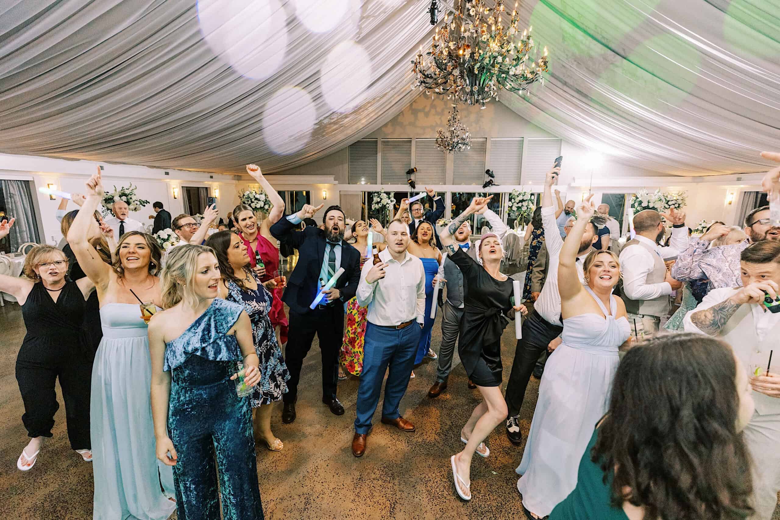 A large group of well-dressed people enthusiastically dancing and celebrating together at an indoor Lake House Inn Wedding, surrounded by decorative lighting and elegant chandeliers.