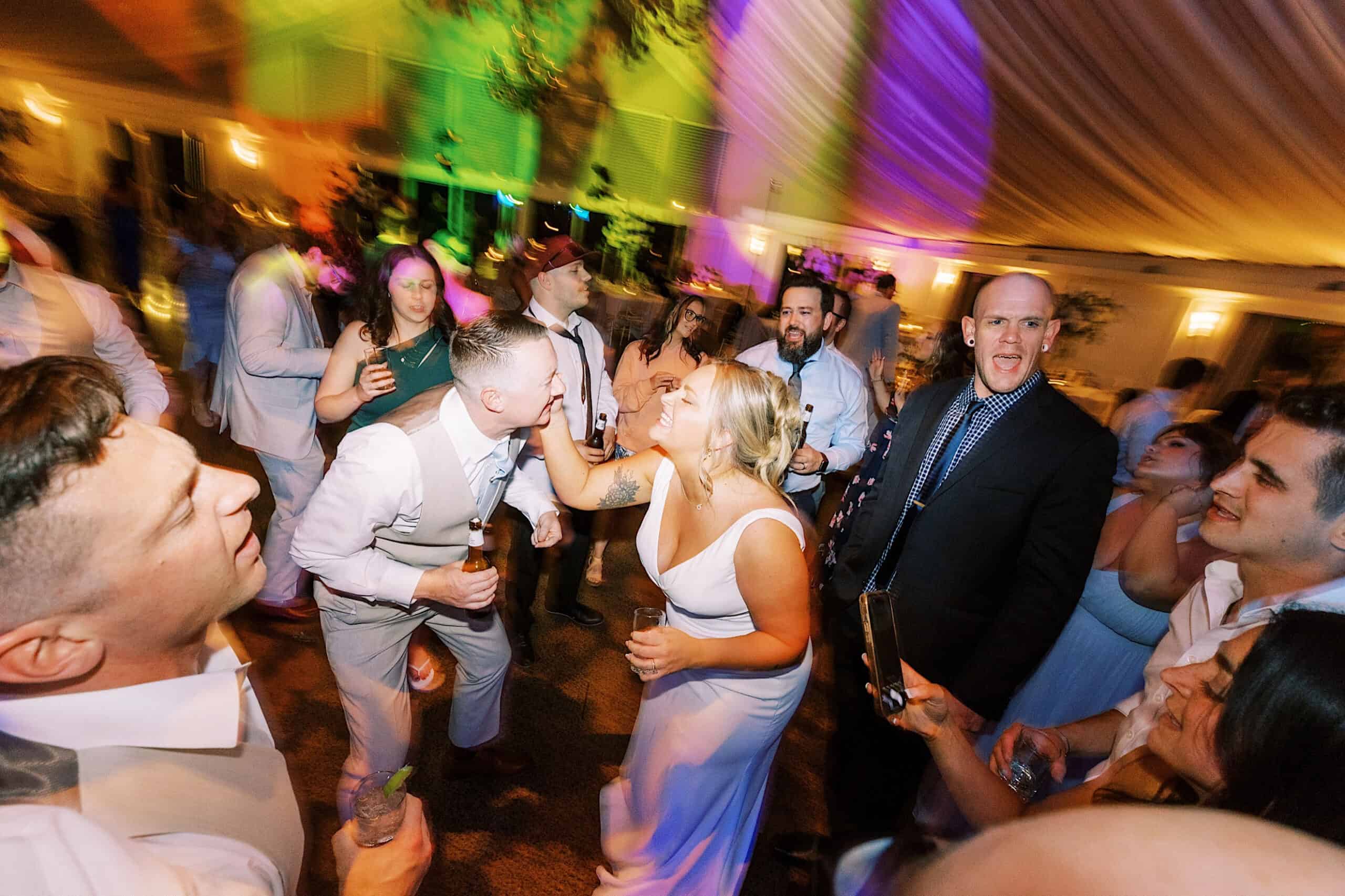 A group of people dance and celebrate at a Lake House Inn Wedding reception, with colorful lights illuminating the scene. The bride feeds the groom as guests smile and laugh around them.