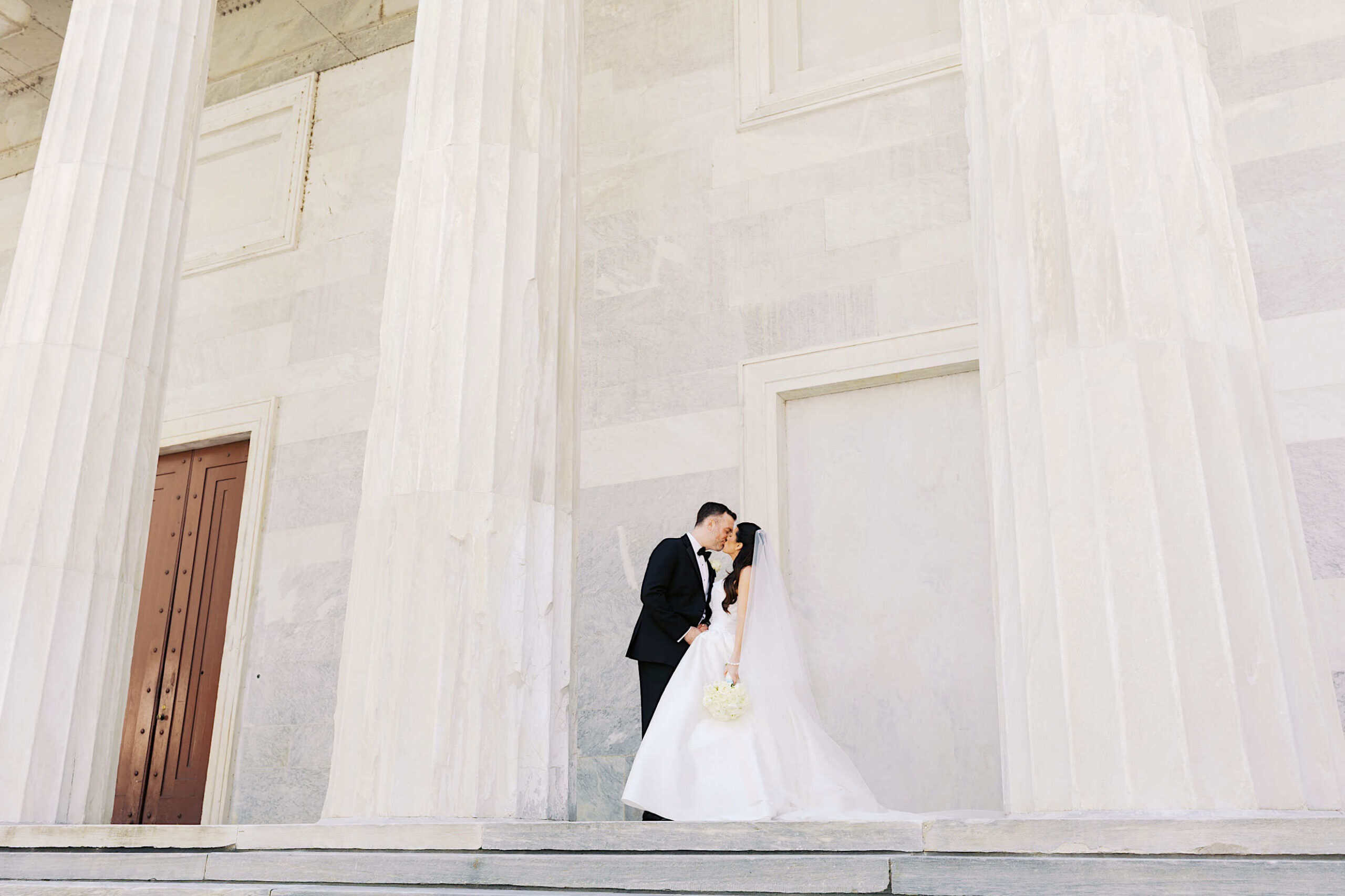A bride and groom stand close together on stone steps in front of a large, white building with tall columns, capturing the elegance of a Classic Wedding at Ballroom at the Ben.