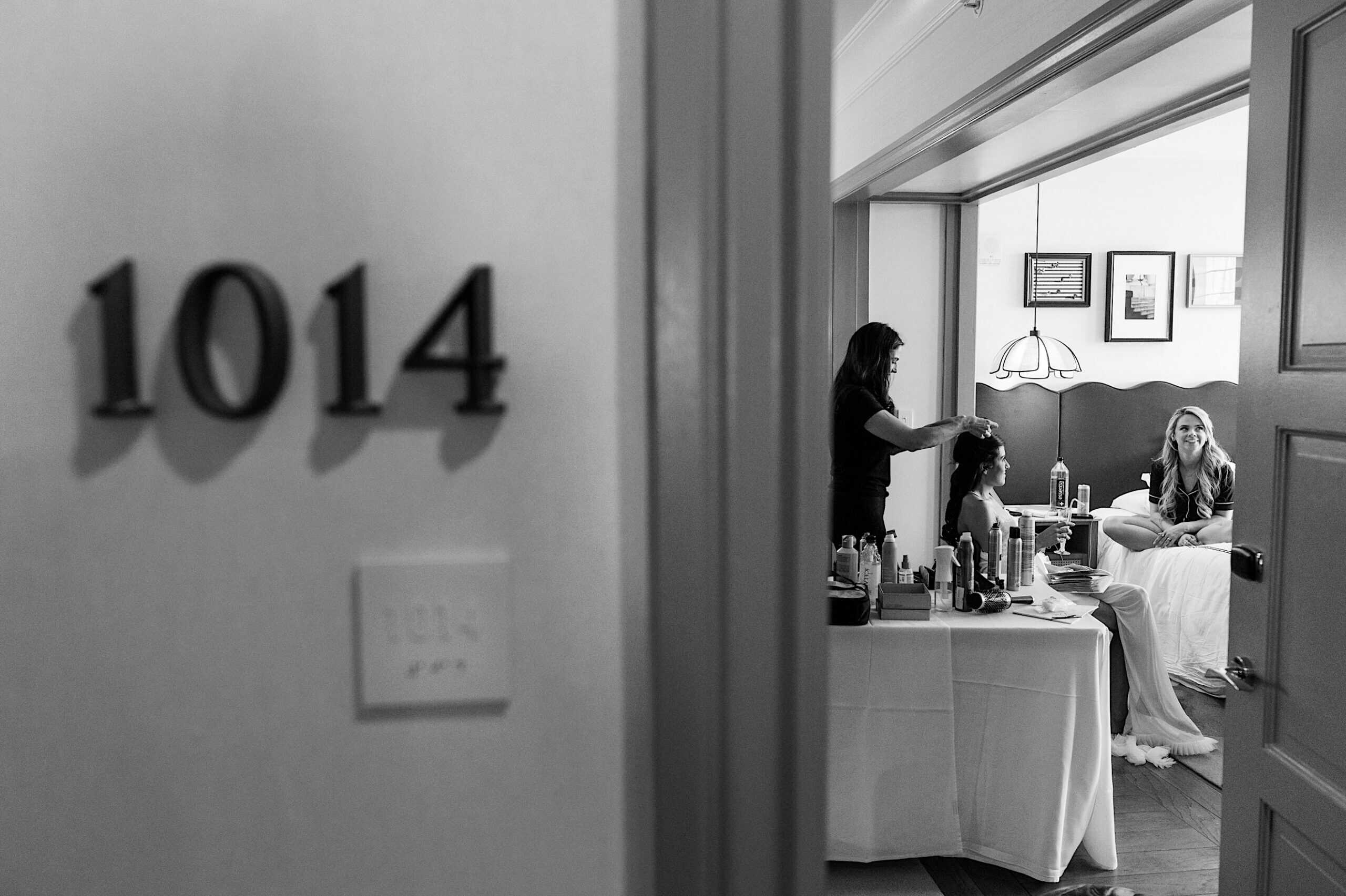 Black and white photo of hotel room 1014, with two women inside, one styling the other's hair while a third woman sits on the bed watching—capturing a quiet moment before a Classic Wedding at Ballroom at the Ben.