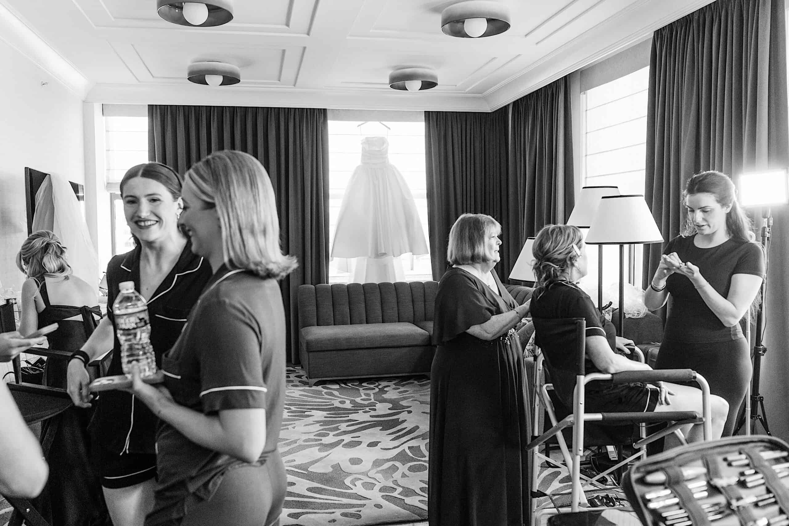 A group of women in a hotel room prepare for a Classic Wedding at Ballroom at the Ben, some seated getting makeup and hair done, with a wedding dress hanging in the window background.