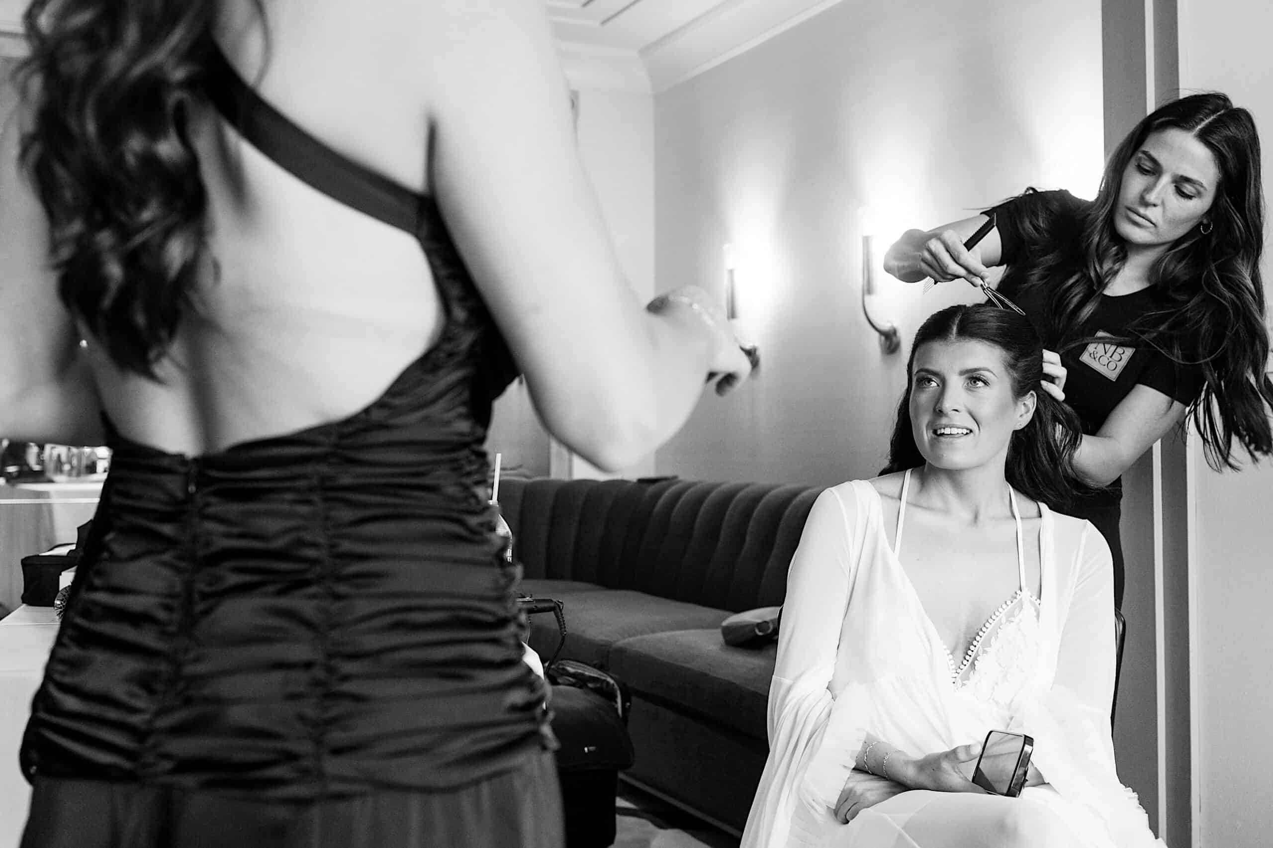 A woman sits on a chair in a robe while a stylist does her hair; another woman in a dress stands nearby holding hairstyling tools, preparing for the Classic Wedding at Ballroom at the Ben.