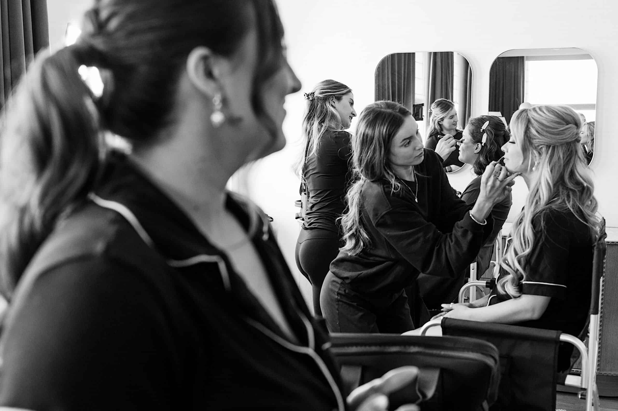 A makeup artist applies lipstick to a seated woman in front of a mirror, preparing her for a classic wedding at Ballroom at the Ben, while another woman stands in the foreground.