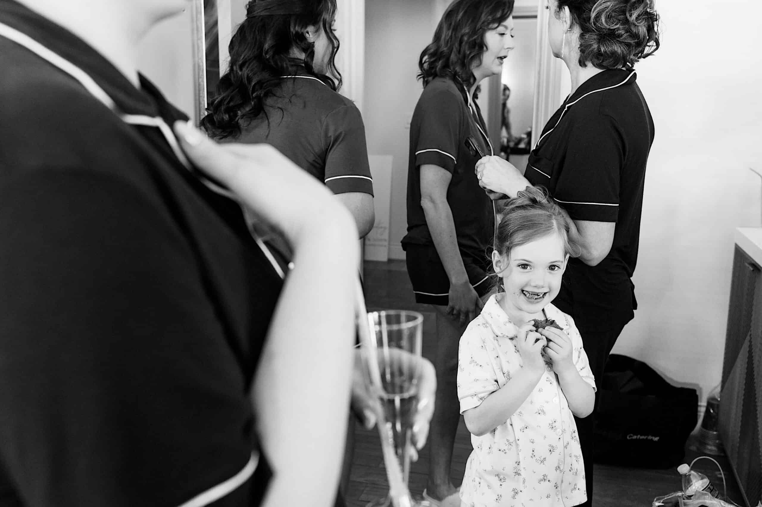 A young girl stands smiling in the foreground while several women, all in pajamas, prepare for a Classic Wedding at Ballroom at the Ben, one holding a glass of champagne.