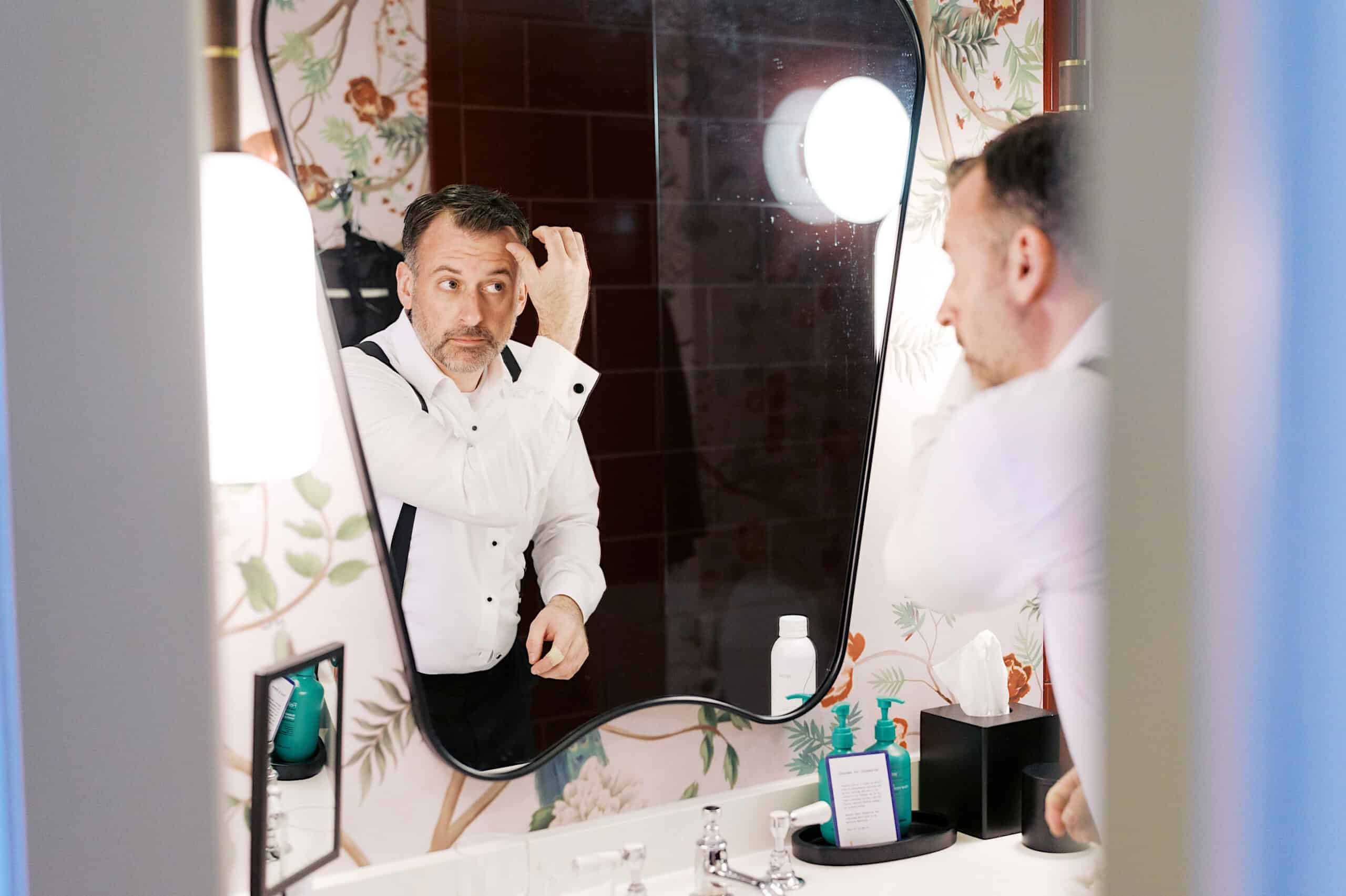 A man in formal attire stands at a bathroom sink, adjusting his hair in front of a mirror with floral wallpaper, preparing for a Classic Wedding at Ballroom at the Ben.