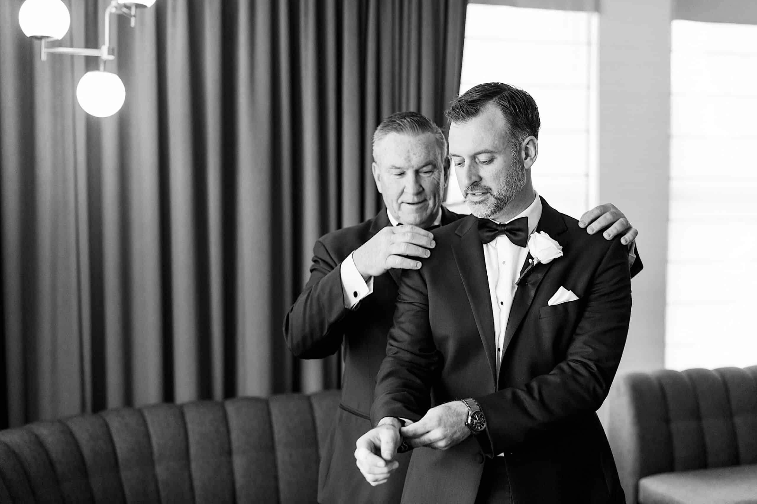 Two men in formal suits stand together; one helps the other adjust his jacket in a room with curtains and a sofa. Black and white photo capturing a Classic Wedding at Ballroom at the Ben.