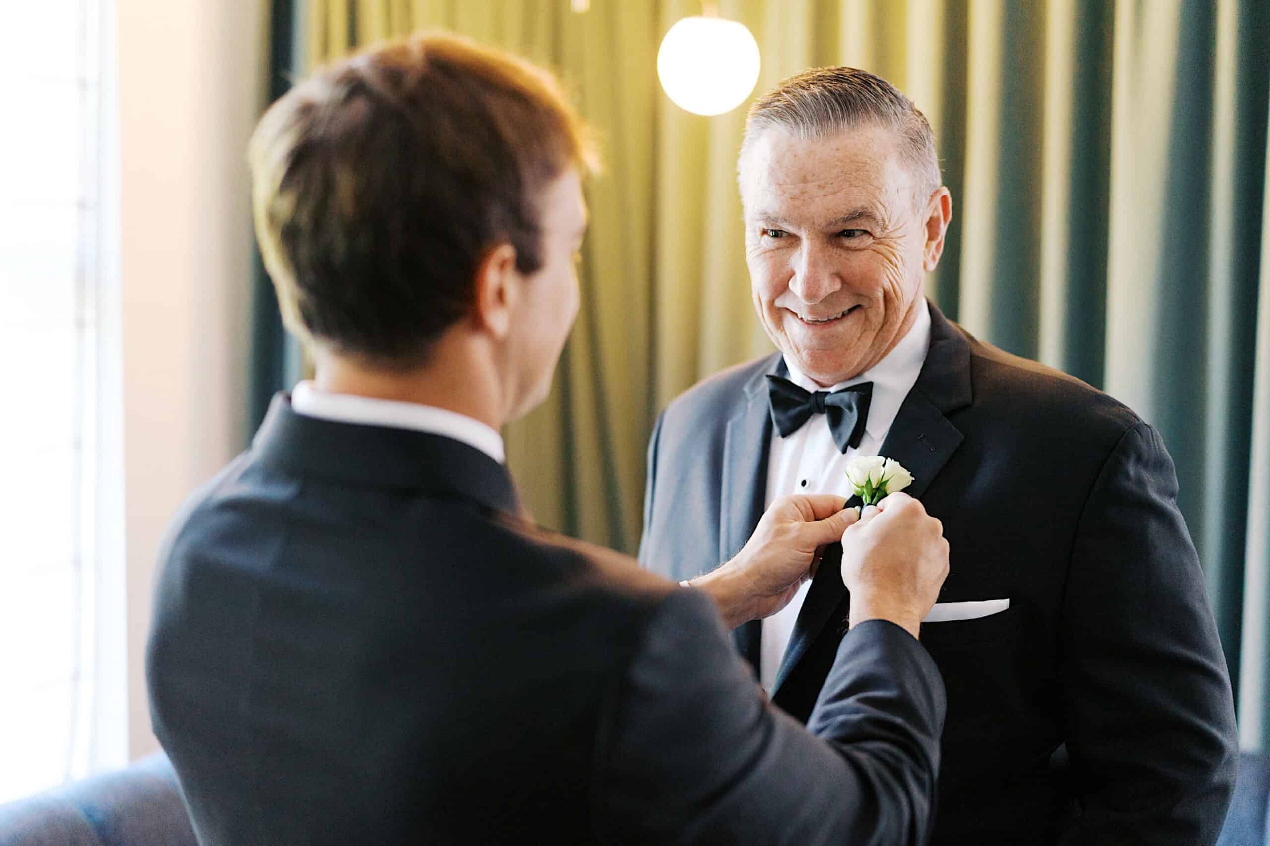 A man in a suit pins a boutonniere onto an older man in a tuxedo, both smiling, at a Classic Wedding at Ballroom at the Ben. They are indoors with green curtains and a round light in the background.