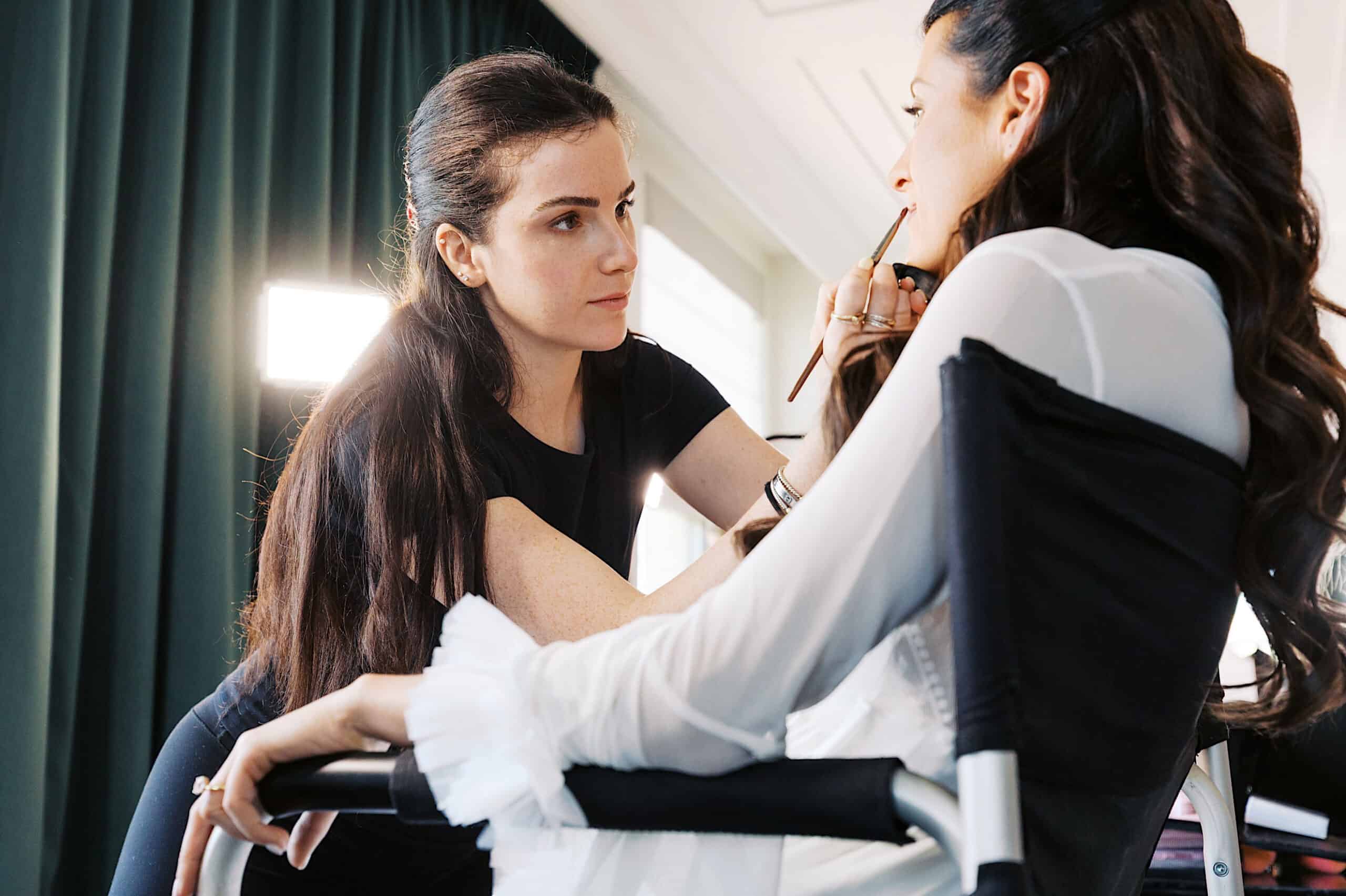 A makeup artist applies lipstick to a seated woman with long hair, dressed in a white top, preparing her for a Classic Wedding at Ballroom at the Ben in a well-lit indoor setting.