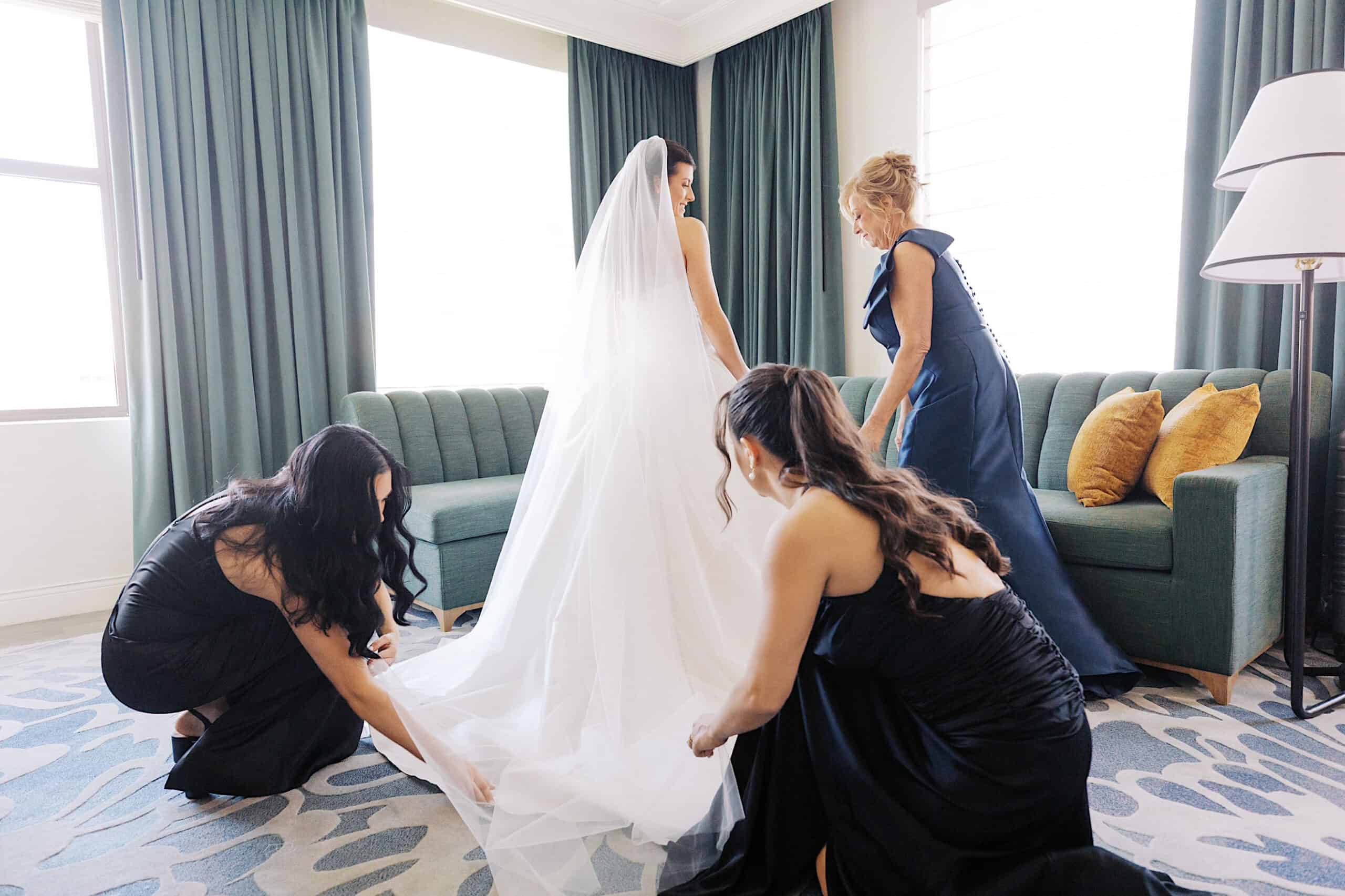 A bride in a white gown stands in a room as two women adjust her train and another woman in a navy dress stands nearby, preparing for a classic wedding at Ballroom at the Ben.