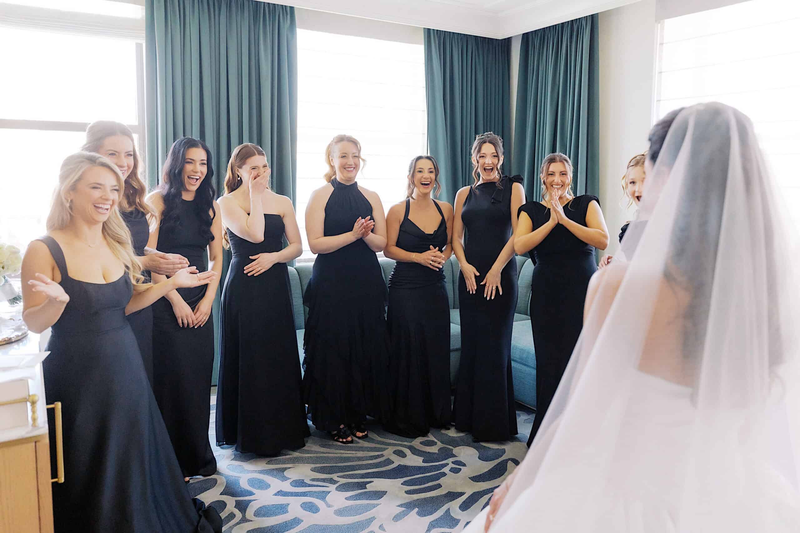 A group of bridesmaids in black dresses stand in a room at the Classic Wedding at Ballroom at the Ben, smiling and reacting with excitement as they look at a bride in a white gown and veil.