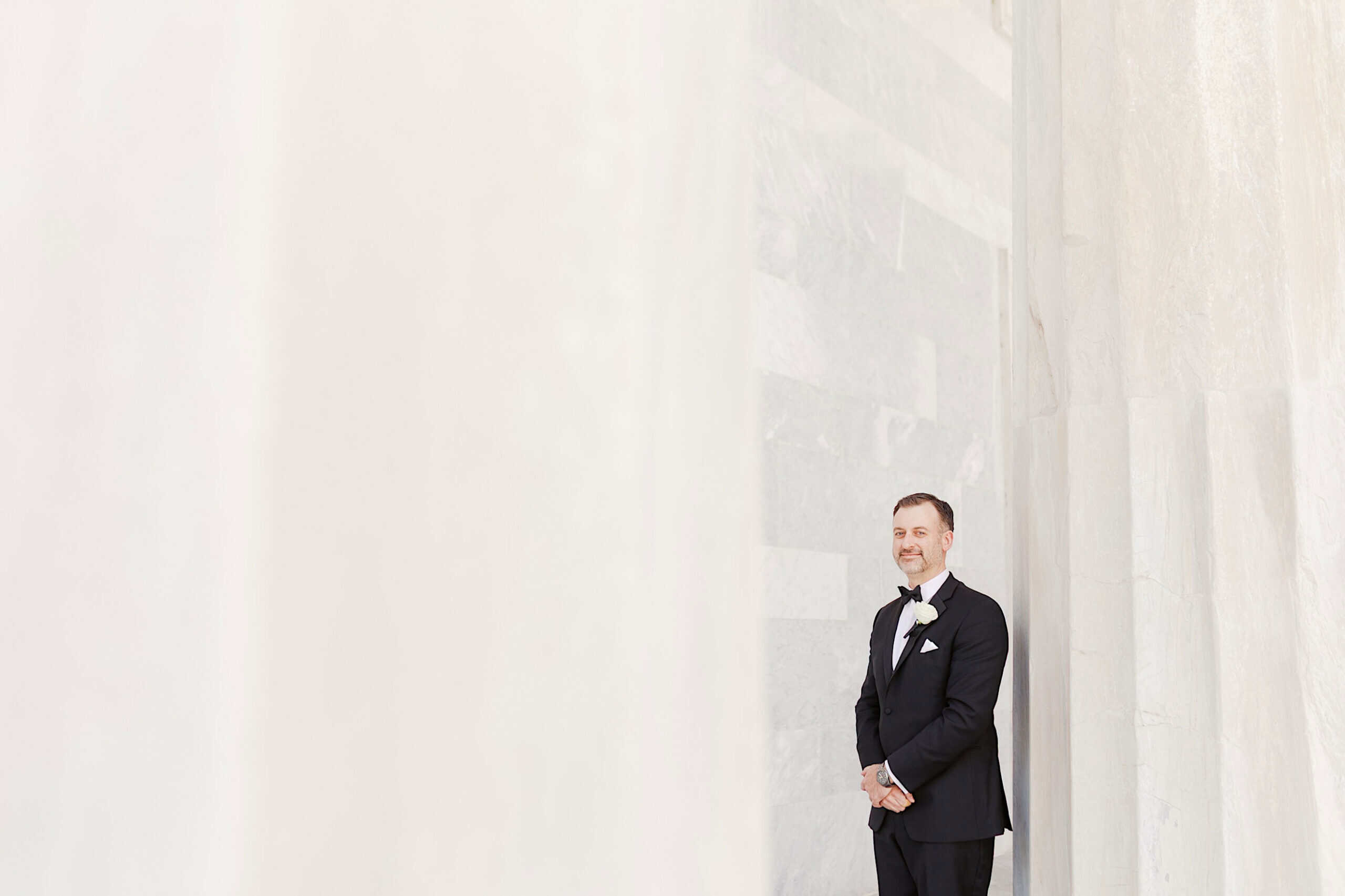 A man in a black tuxedo stands and smiles near tall, white marble columns with bright light illuminating the scene at a Classic Wedding at Ballroom at the Ben.