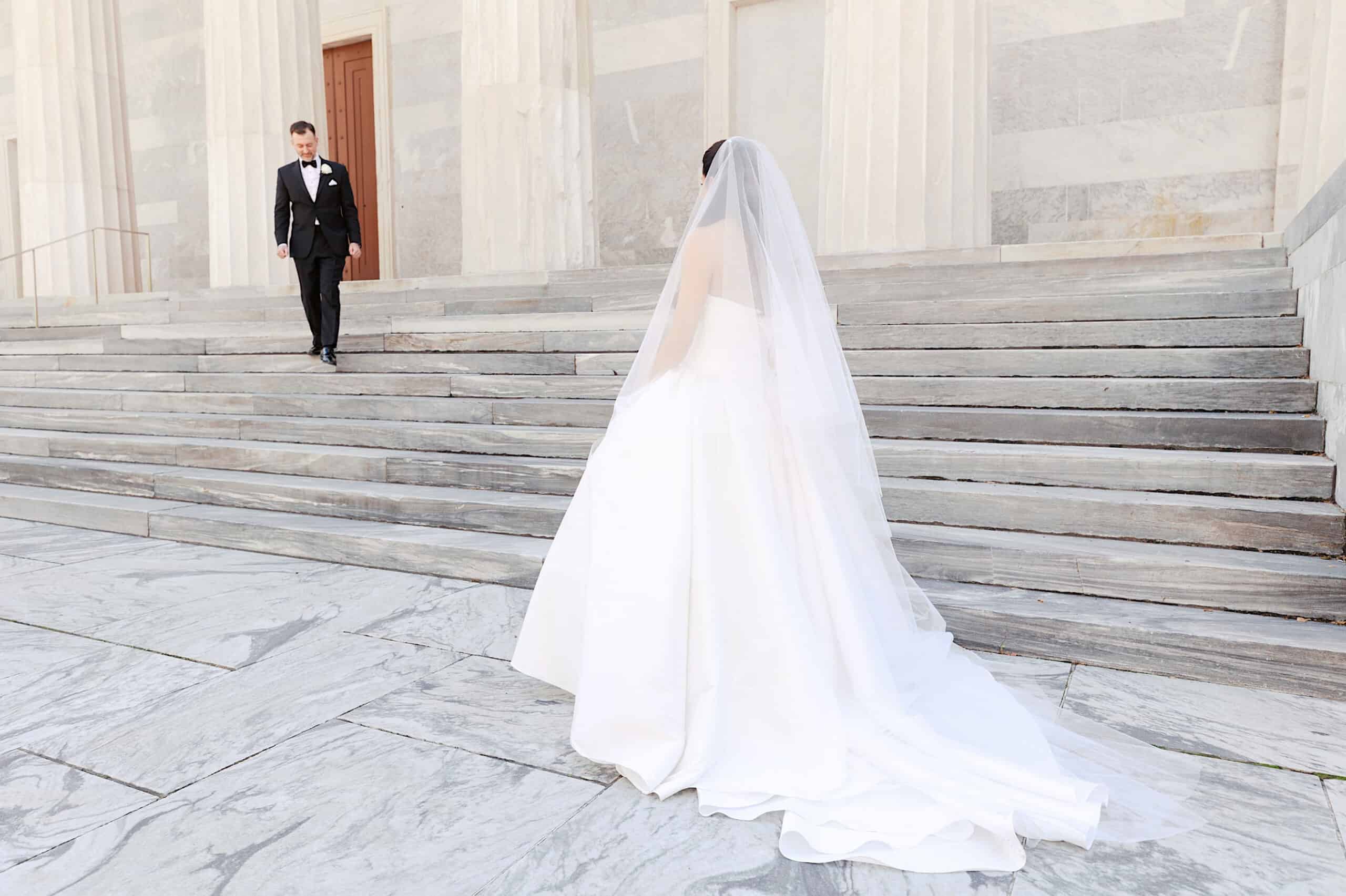 A bride in a white gown and veil stands at the bottom of stone steps, facing her groom in a tuxedo as he walks down toward her—a timeless moment from their Classic Wedding at Ballroom at the Ben.