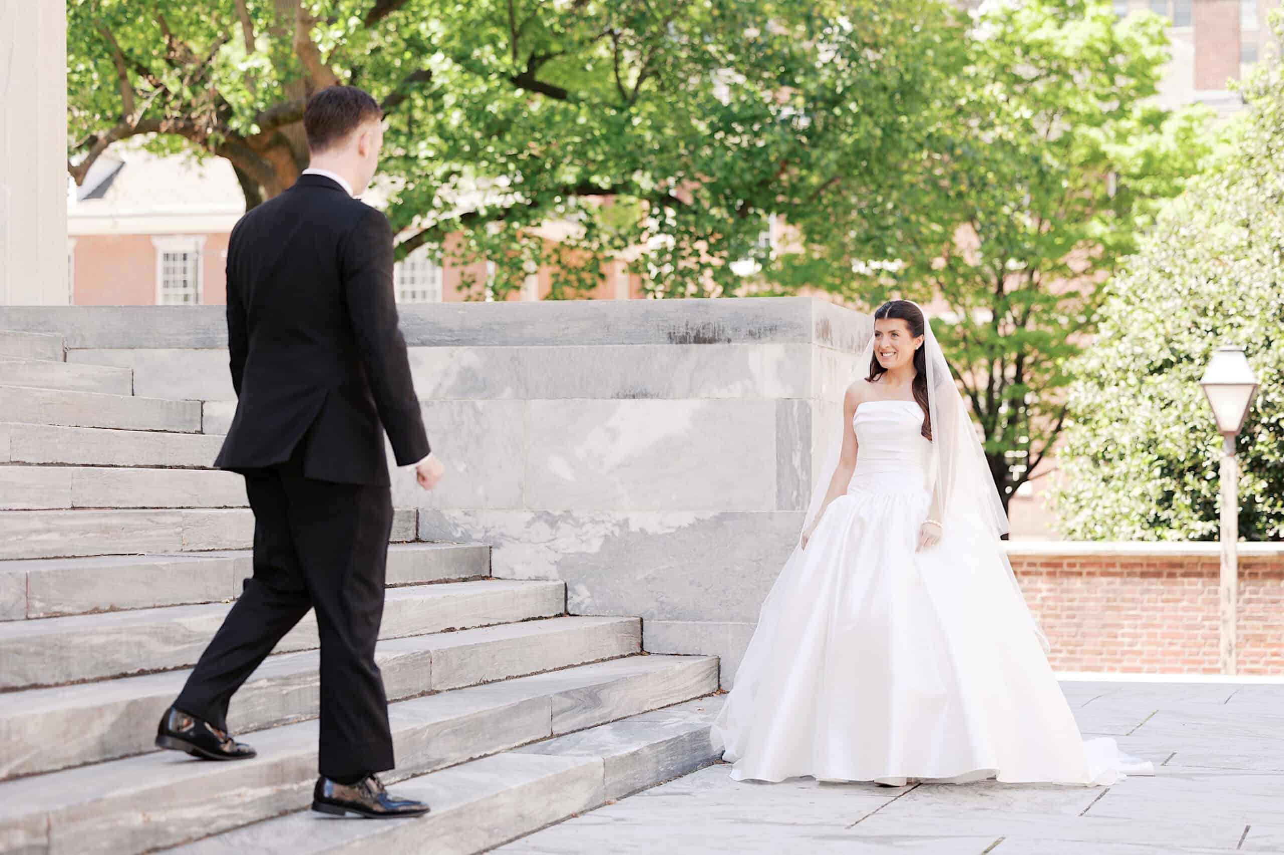 A bride in a white gown smiles at her groom in a black suit as he walks down stone steps outdoors, with trees and sunlight in the background, capturing a timeless moment from their Classic Wedding at Ballroom at the Ben.