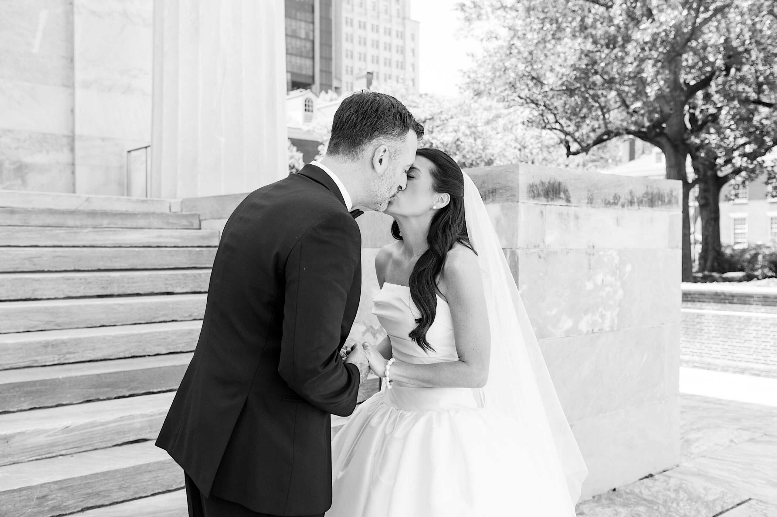 A bride and groom kiss outside on their wedding day near stone steps and a large tree, capturing a timeless moment before their Classic Wedding at Ballroom at the Ben.