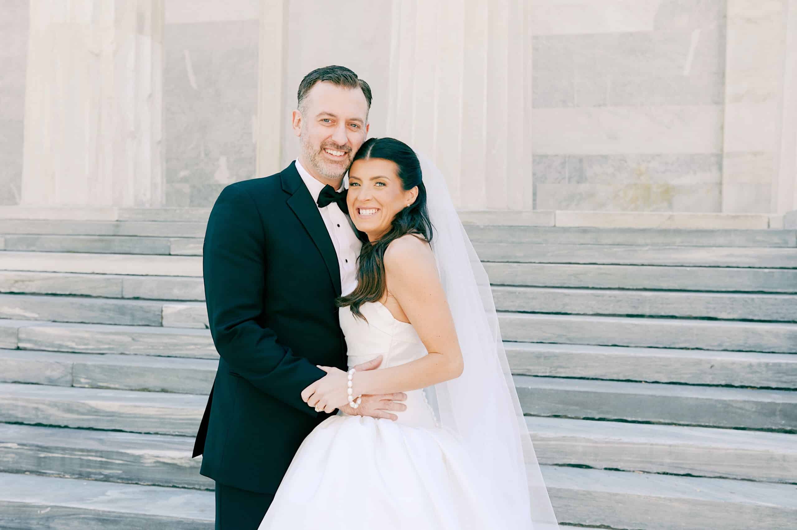 A bride in a white dress and veil stands next to a groom in a black tuxedo, both smiling in front of stone steps at their classic wedding at Ballroom at the Ben.
