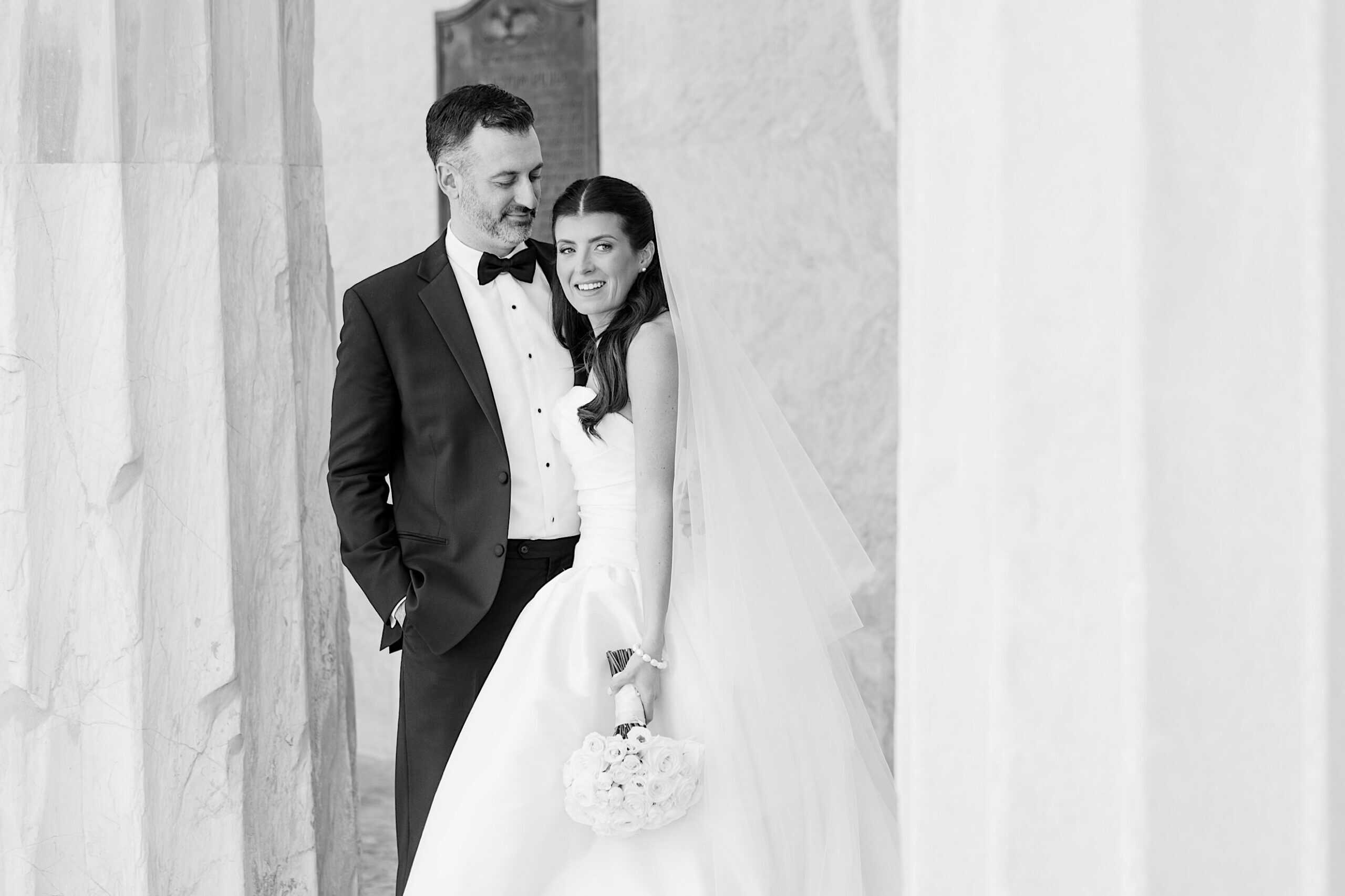 A bride and groom in formal attire stand together, smiling, with marble columns in the background at their Classic Wedding at Ballroom at the Ben. The bride holds a bouquet and wears a veil.