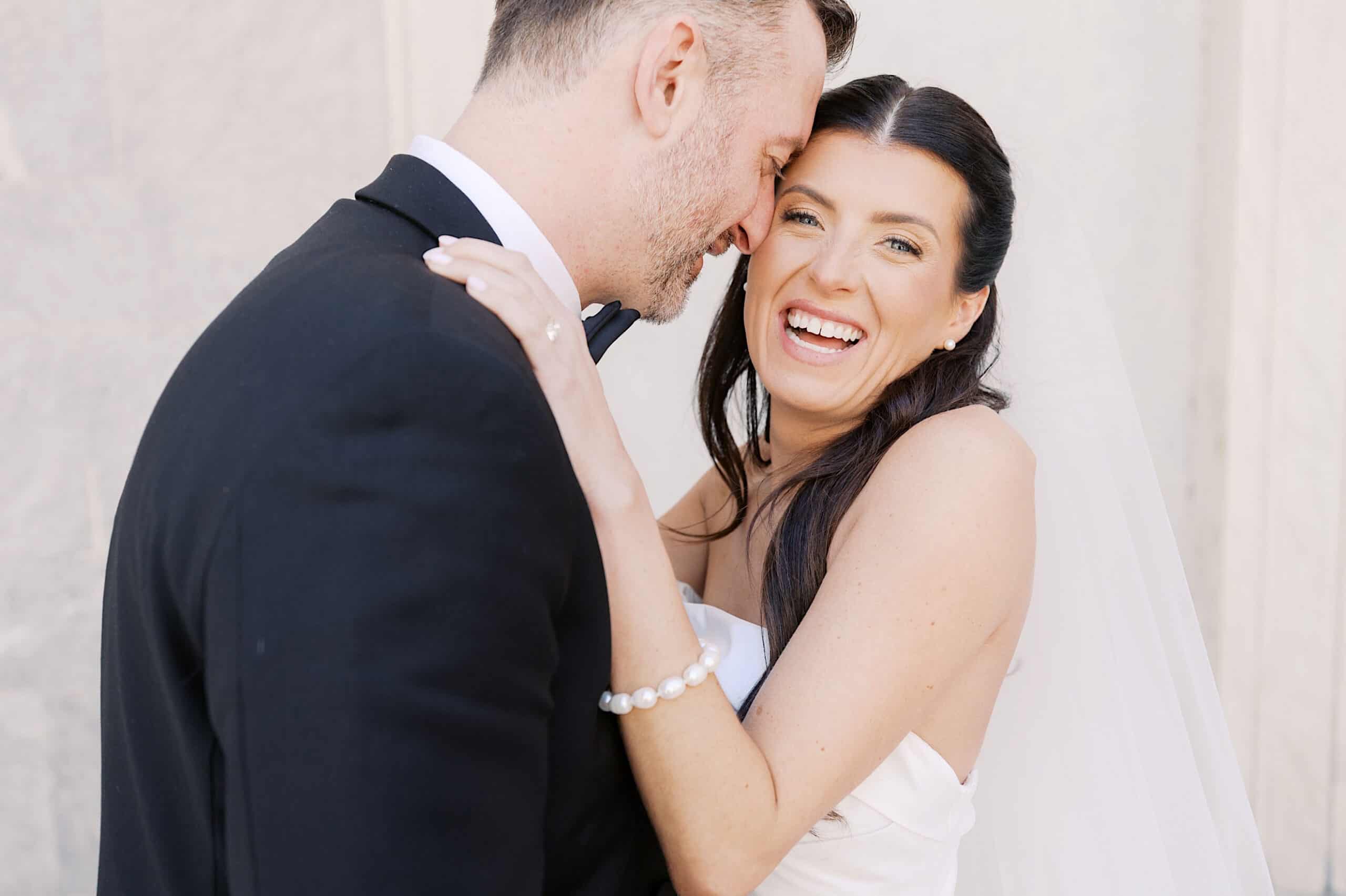 A bride in a white dress and veil smiles while embracing a groom in a black suit. They stand close together in front of a light-colored background, capturing the elegance of a Classic Wedding at Ballroom at the Ben.