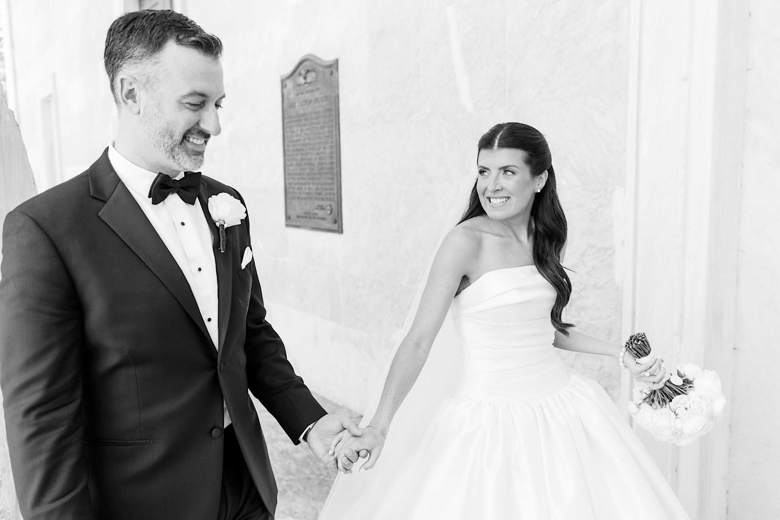 A bride in a white gown holds a bouquet and smiles at her groom in a black tuxedo as they hold hands and walk outside after their Classic Wedding at Ballroom at the Ben.