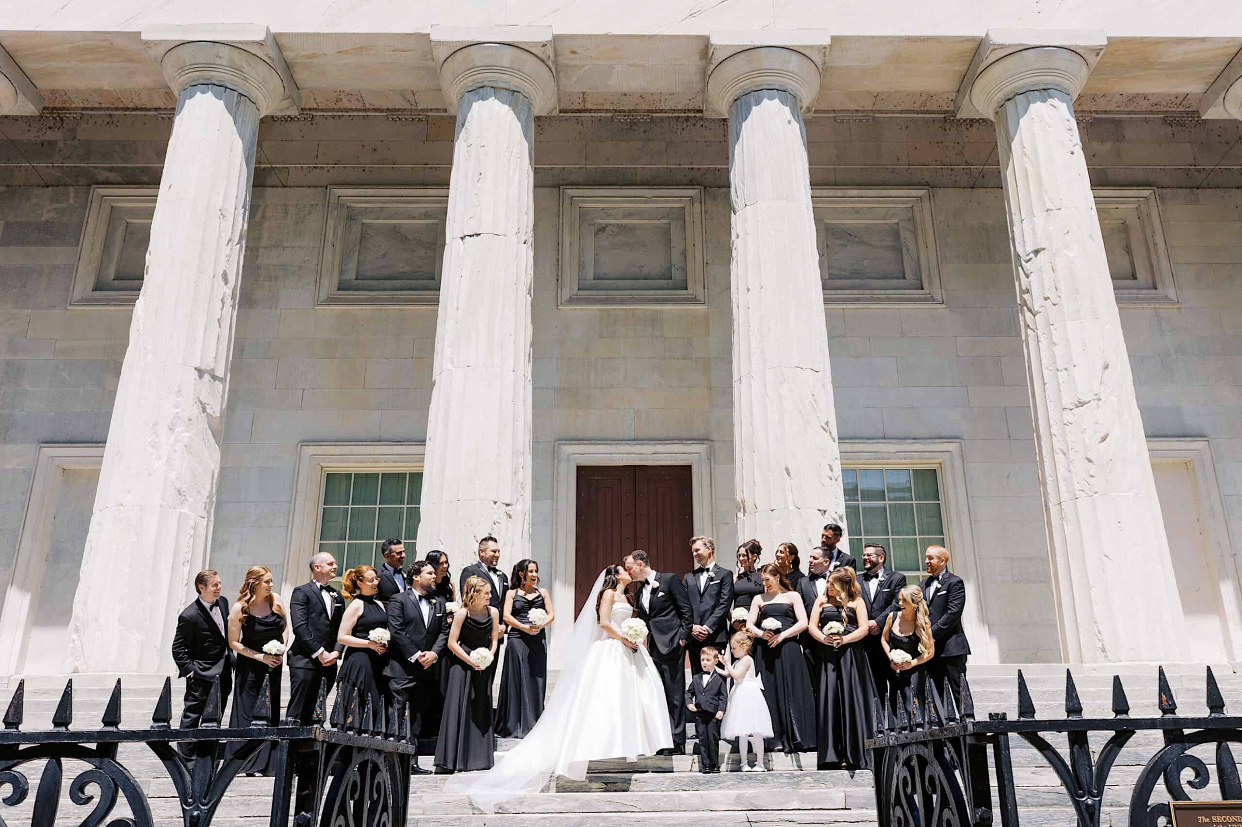 A Classic Wedding at Ballroom at the Ben: A wedding party stands on the steps of a neoclassical building with tall columns, with the bride and groom in the center and bridesmaids and groomsmen on either side.