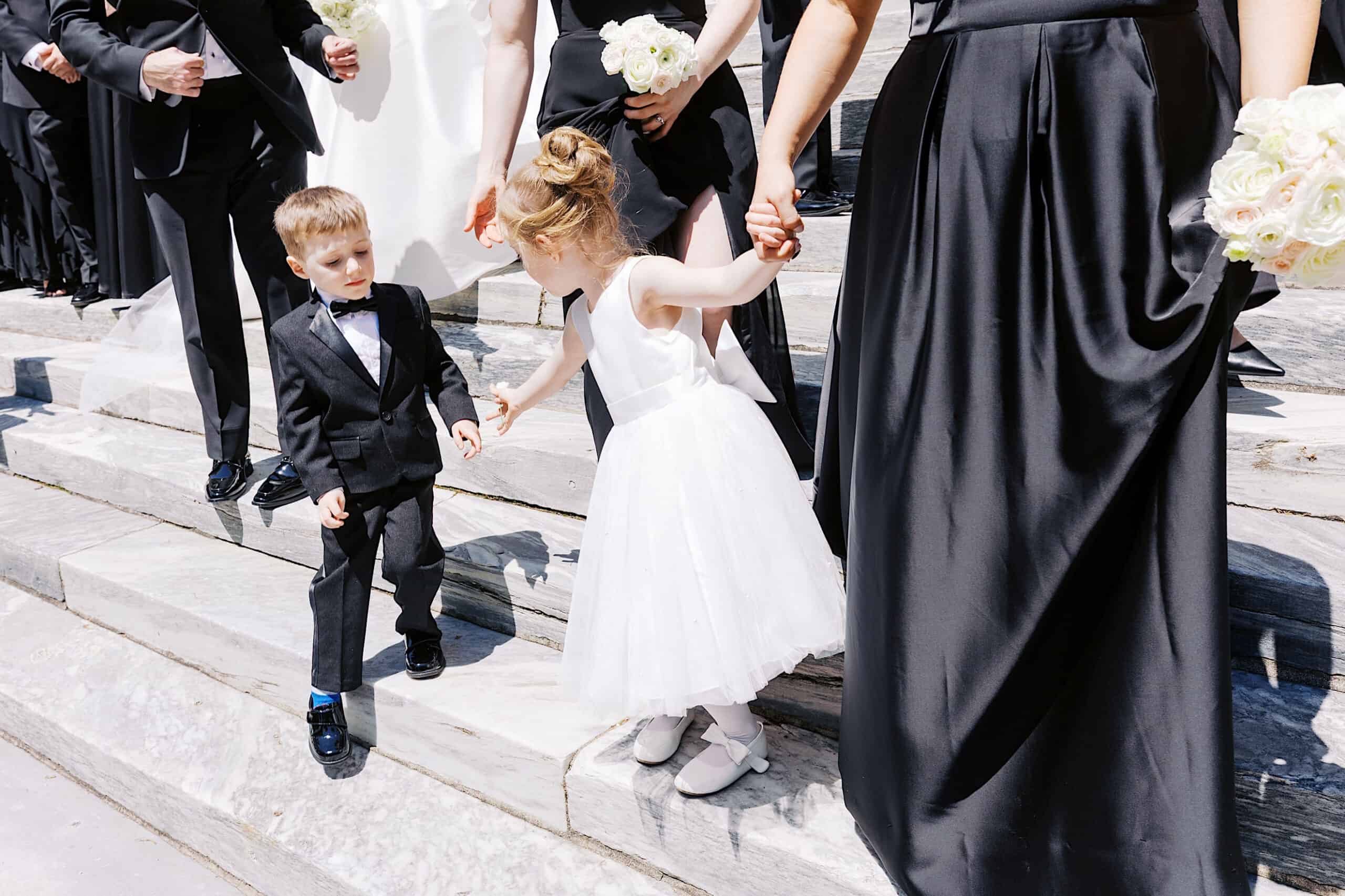 A young boy in a suit and a girl in a white dress hold hands on stone steps, surrounded by adults in black formal attire with white bouquets at this Classic Wedding at Ballroom at the Ben.