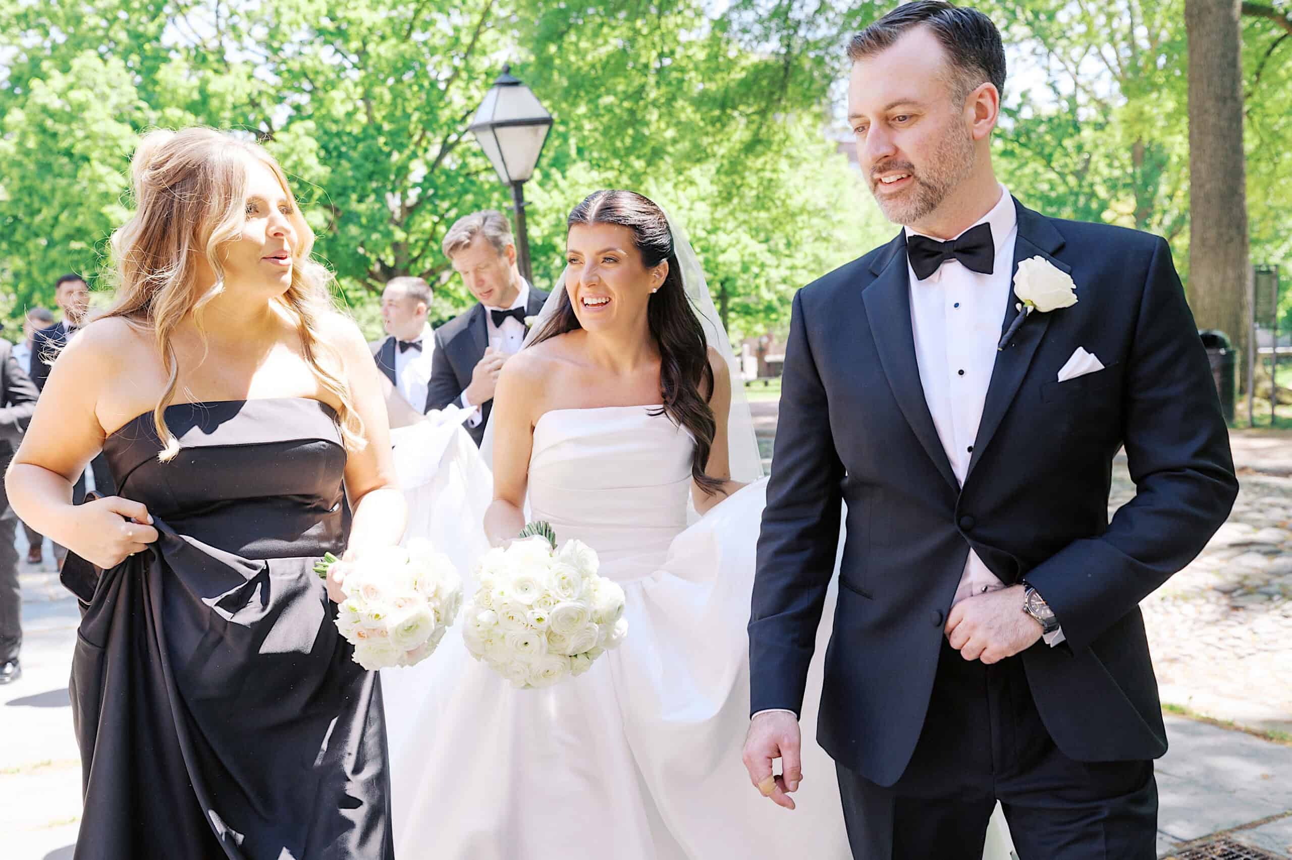 A bride in a white dress walks outdoors with a bridesmaid in a black dress and a man in a tuxedo, each holding white bouquets—capturing the elegance of a Classic Wedding at Ballroom at the Ben.