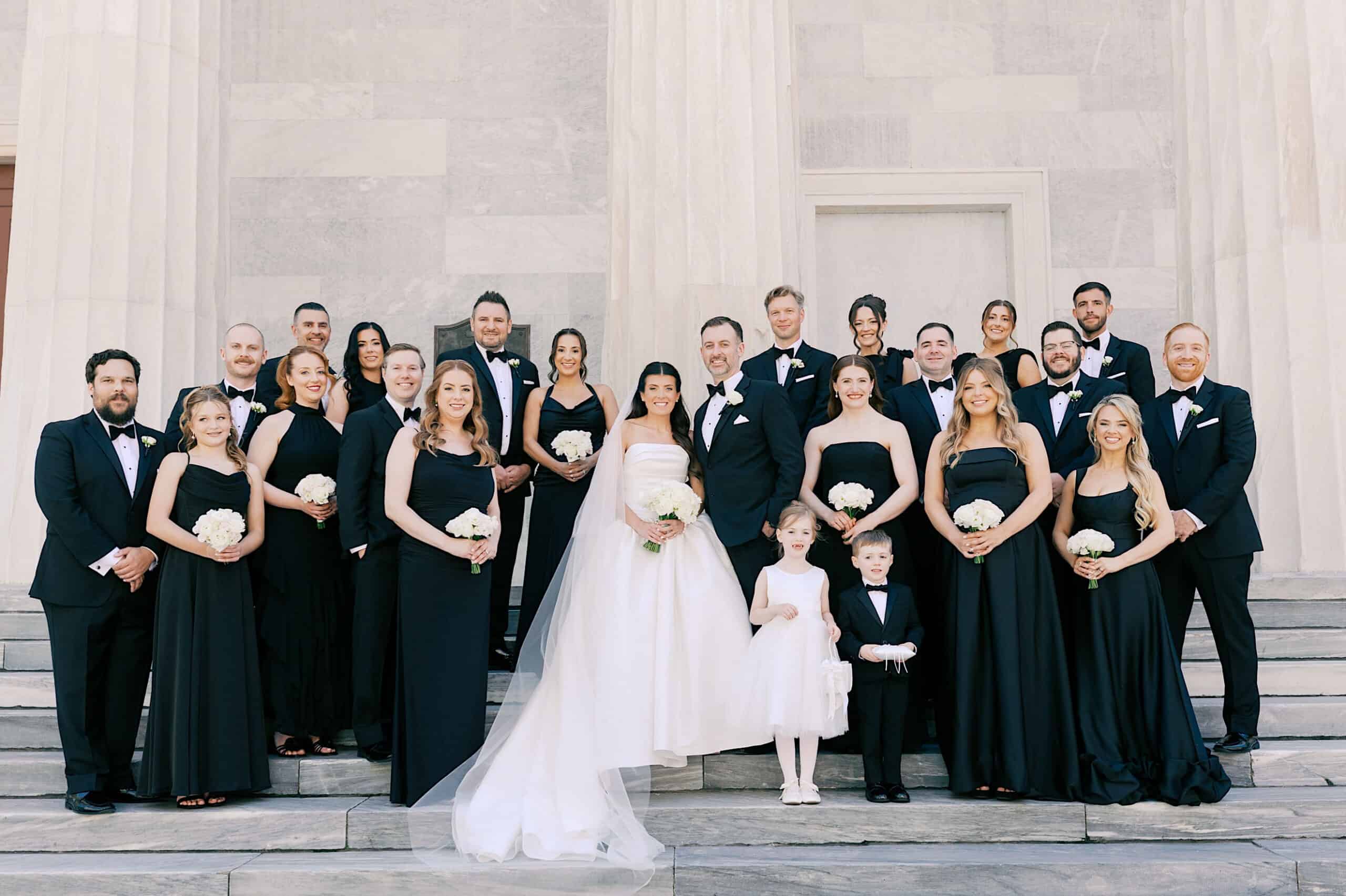 A wedding party poses on stone steps at the Classic Wedding at Ballroom at the Ben; the bride stands in the center in a white gown, surrounded by bridesmaids in black dresses and groomsmen in black suits.