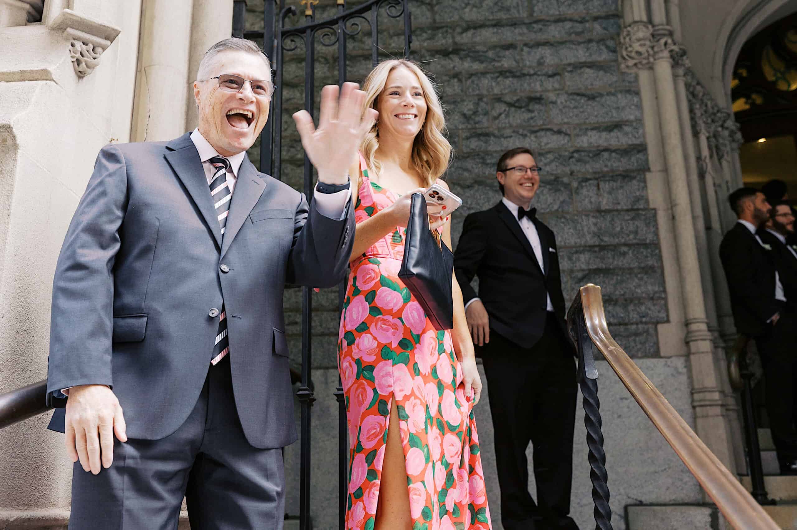 A man in a suit waves and smiles next to a woman in a floral dress outside a stone building, with other guests in formalwear nearby—capturing the joyful spirit of a classic wedding at Ballroom at the Ben.