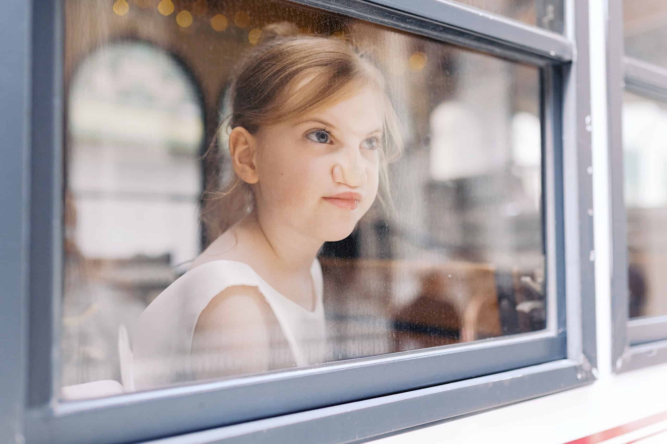 A young girl in a white dress presses her nose and lips against a window, looking outside with a playful expression during a classic wedding at Ballroom at the Ben.