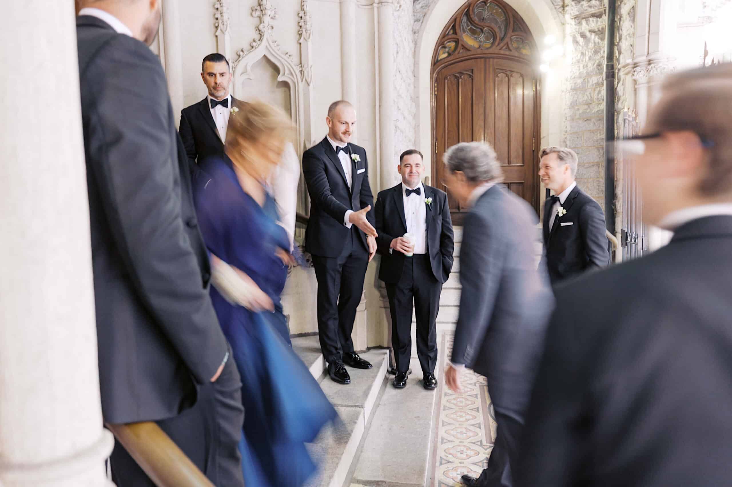 A group of people in formal attire stand and walk on a staircase inside an ornate building with arched doorways and decorative stonework, capturing the elegance of a Classic Wedding at Ballroom at the Ben.