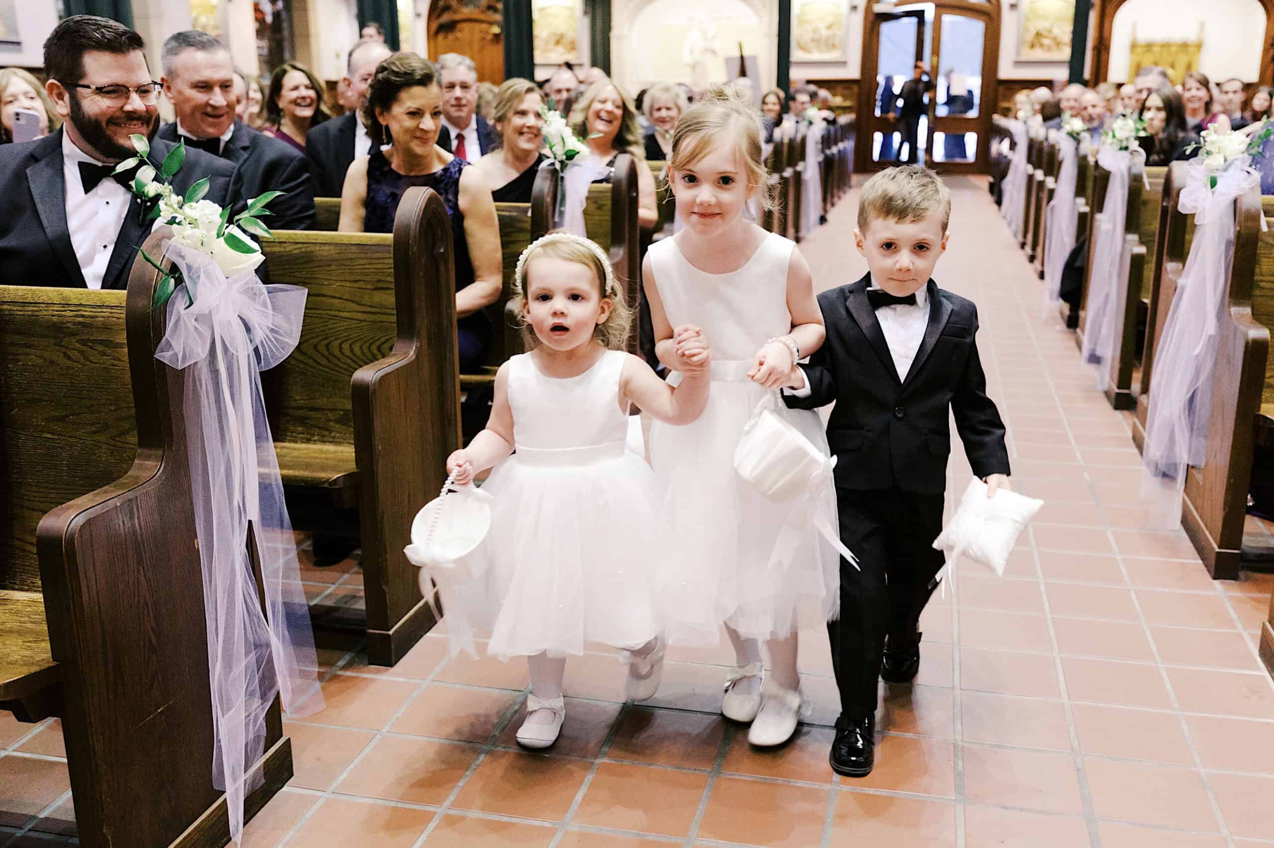 Three young children, two girls in white dresses and a boy in a black suit, walk down the aisle of a church, holding hands, during a Classic Wedding at Ballroom at the Ben.