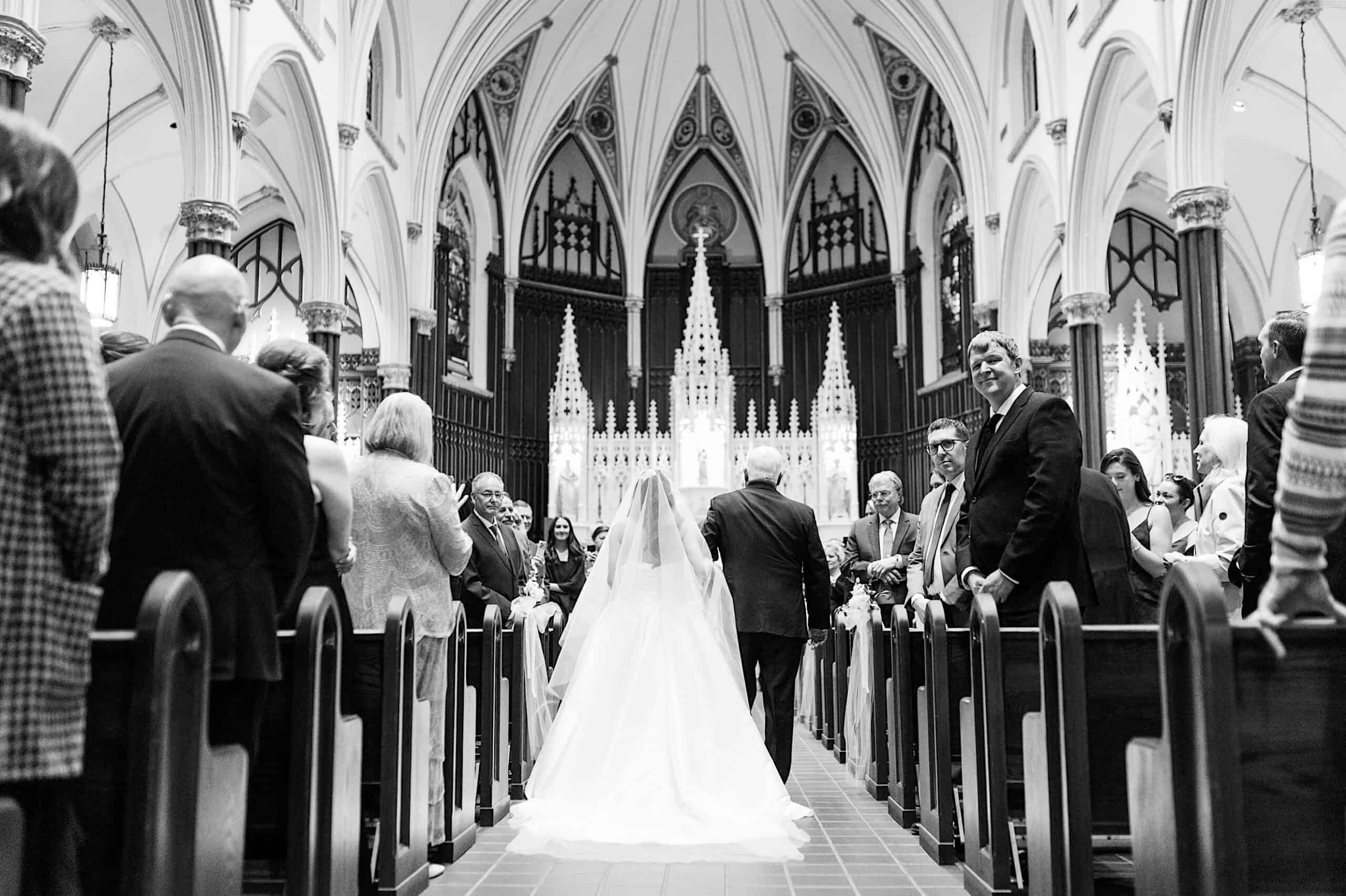 A bride in a white gown walks down the aisle of a grand church, accompanied by an older man, as guests watch from the pews—a scene reminiscent of a Classic Wedding at Ballroom at the Ben.