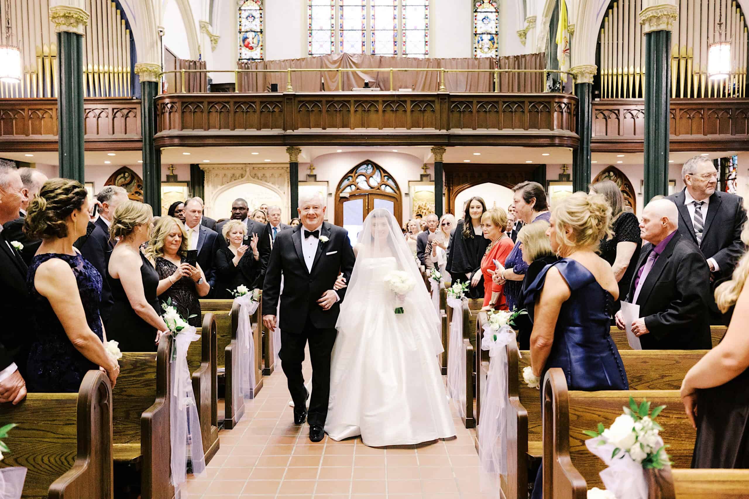 A bride in a white gown walks down the aisle with an older man in a tuxedo, surrounded by guests at a Classic Wedding at Ballroom at the Ben, with the church beautifully decorated for the ceremony.