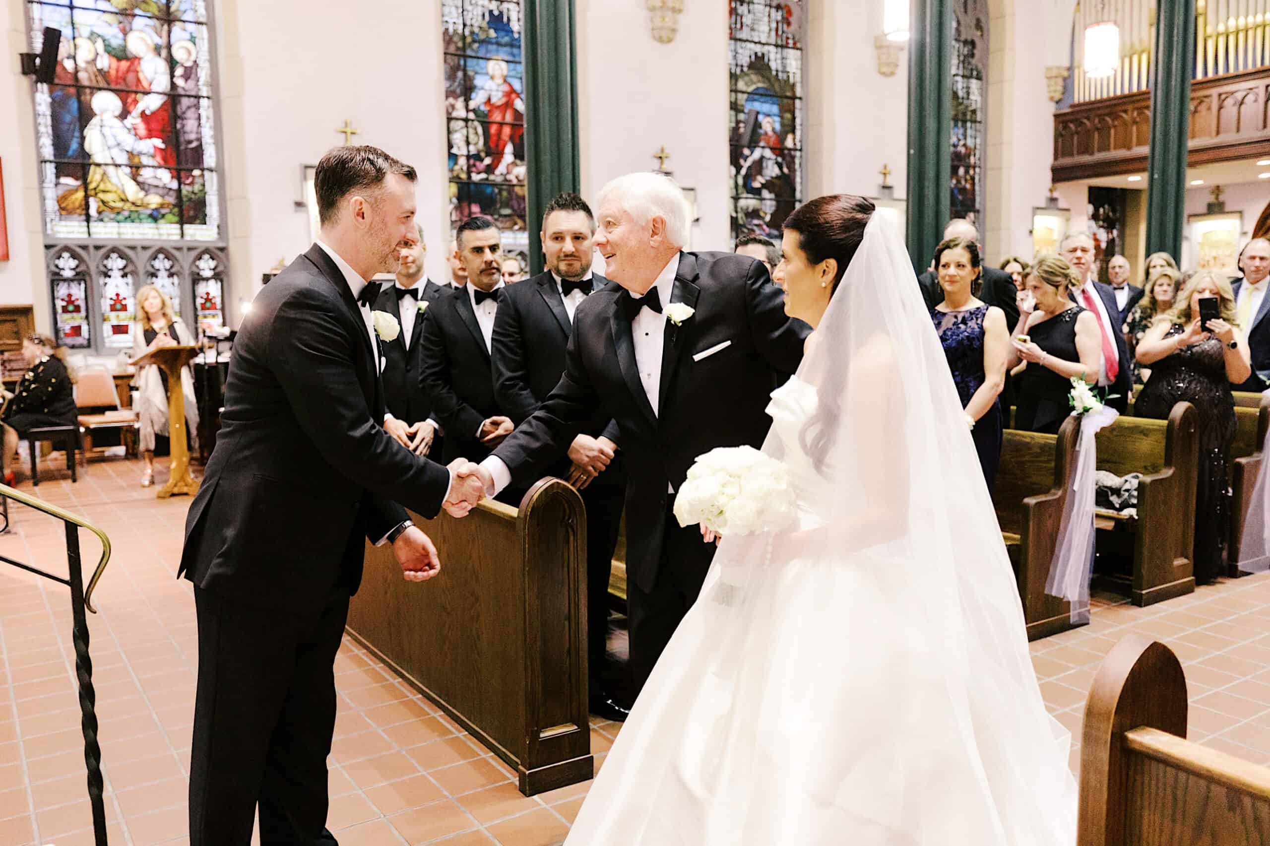 A bride in a white gown stands with her father, who shakes hands with the groom at the altar, as guests look on—capturing the elegance of a Classic Wedding at Ballroom at the Ben.