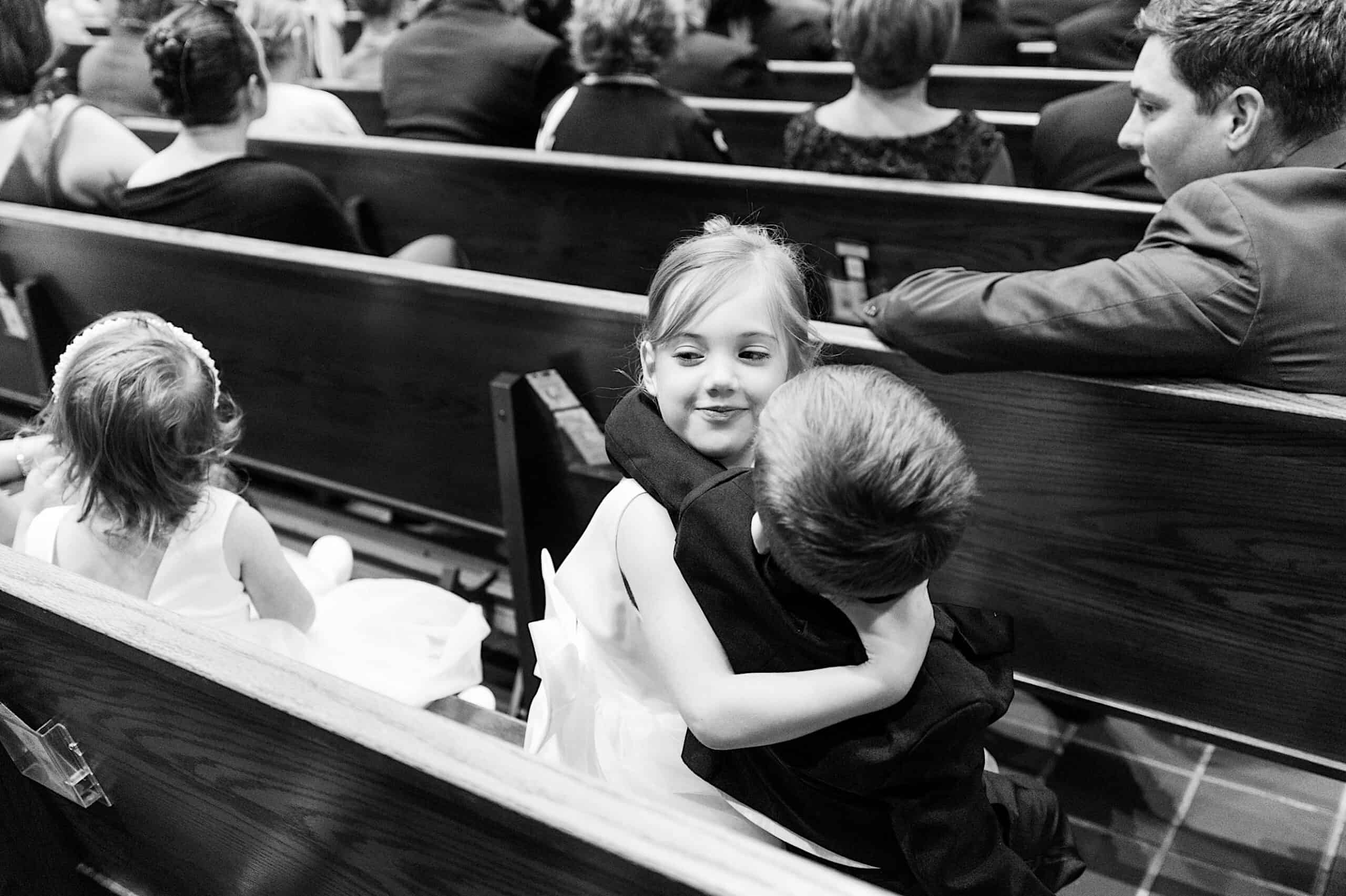 A young girl and boy sit in a church pew, with the girl hugging the boy. Another young girl in a white dress sits nearby. Adults, all guests of a classic wedding at Ballroom at the Ben, are seated in the rows ahead.