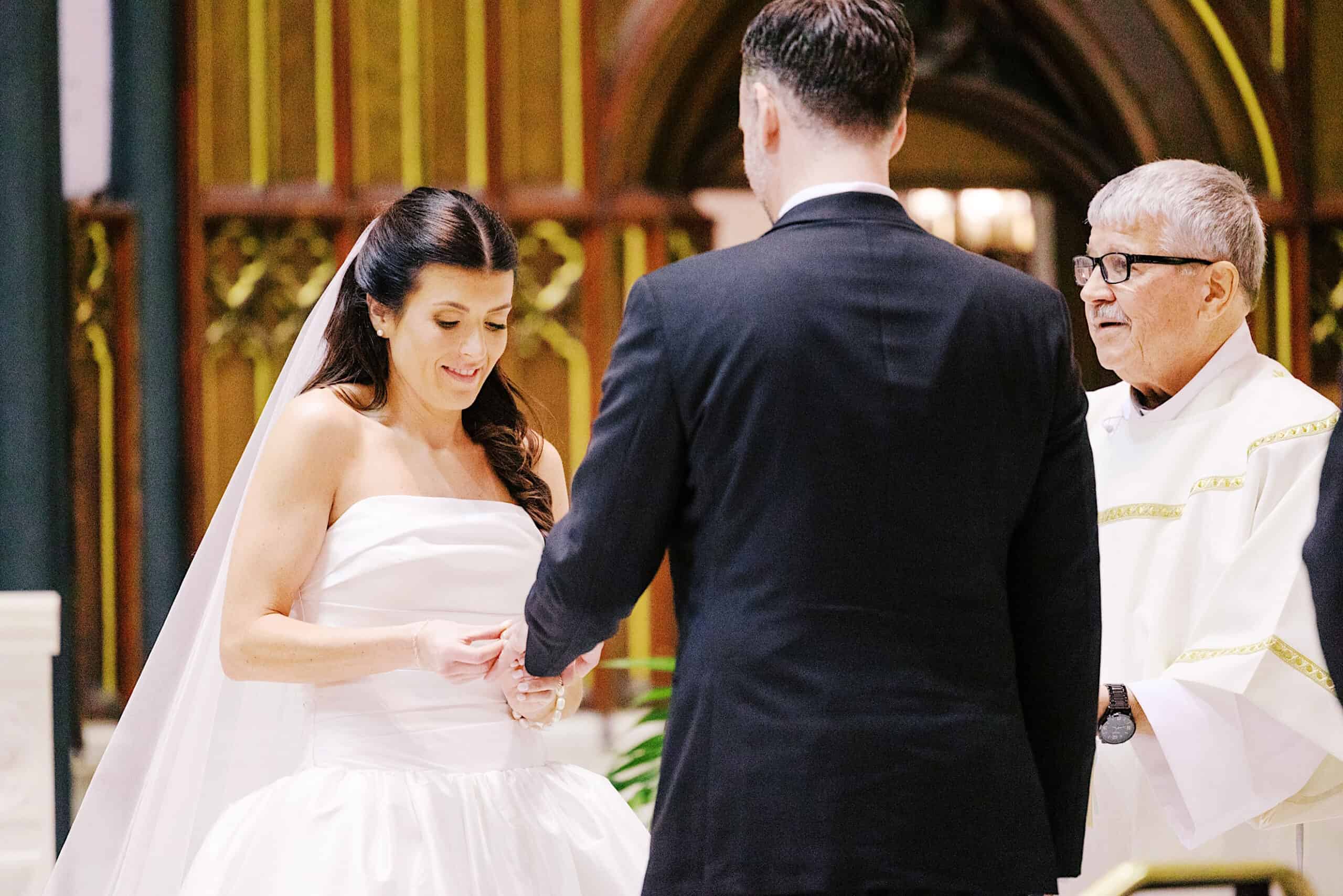 A bride and groom stand facing each other, holding hands, as a priest officiates their classic wedding at Ballroom at the Ben.
